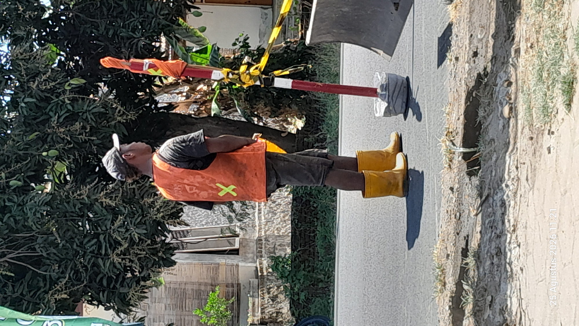 A person wearing a dark t-shirt, dark shorts, a grey baseball cap, an orange safety vest with a green 'X' symbol, and yellow rubber boots stands on an asphalt road. The person is facing away from the camera, with their head turned to the right. A red and white pole with yellow caution tape wrapped around it stands to the right of the person, anchored in a grey base. Trees with green leaves and some fruit are visible in the background, along with a building. The scene appears to be in Jomblang, Indonesia, as indicated by the timestamp "25 Agustus 2026 11:21". The lighting suggests it is daytime, likely sunny.