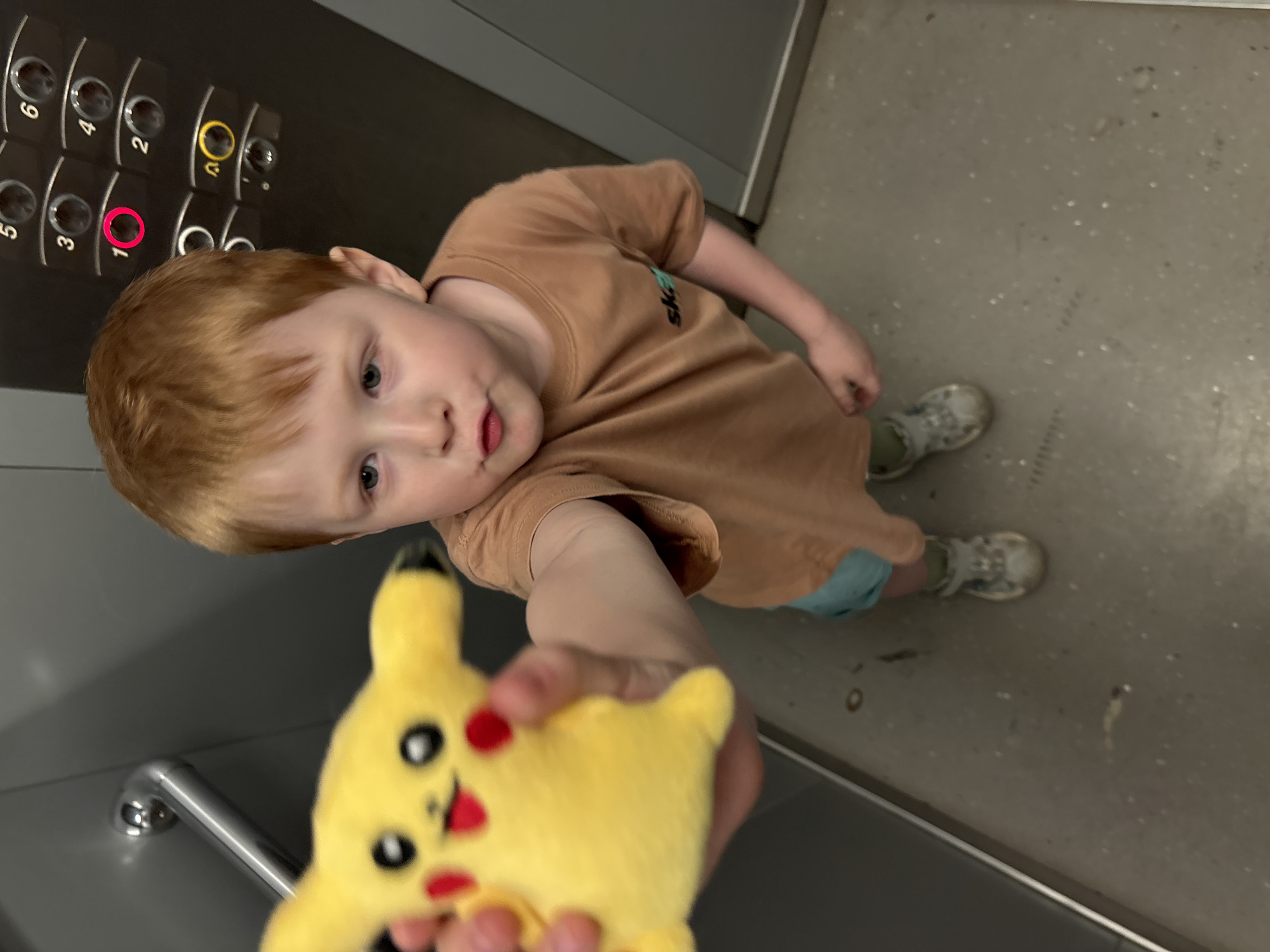 A high-angle shot shows a young, light-skinned, red-haired child in a brown t-shirt, holding a yellow Pikachu plush toy in front of them, seemingly presenting it to the camera. The scene is inside an elevator. Elevator buttons numbered 1-6 are visible in the upper left of the frame, with the '1' button highlighted in red and '0' in yellow. The elevator walls are grey, and the floor is a speckled grey. The child is wearing light-colored sneakers and a blue shirt. The location cannot be determined, especially not as the city of Izhevsk, Russia, with the provided information.