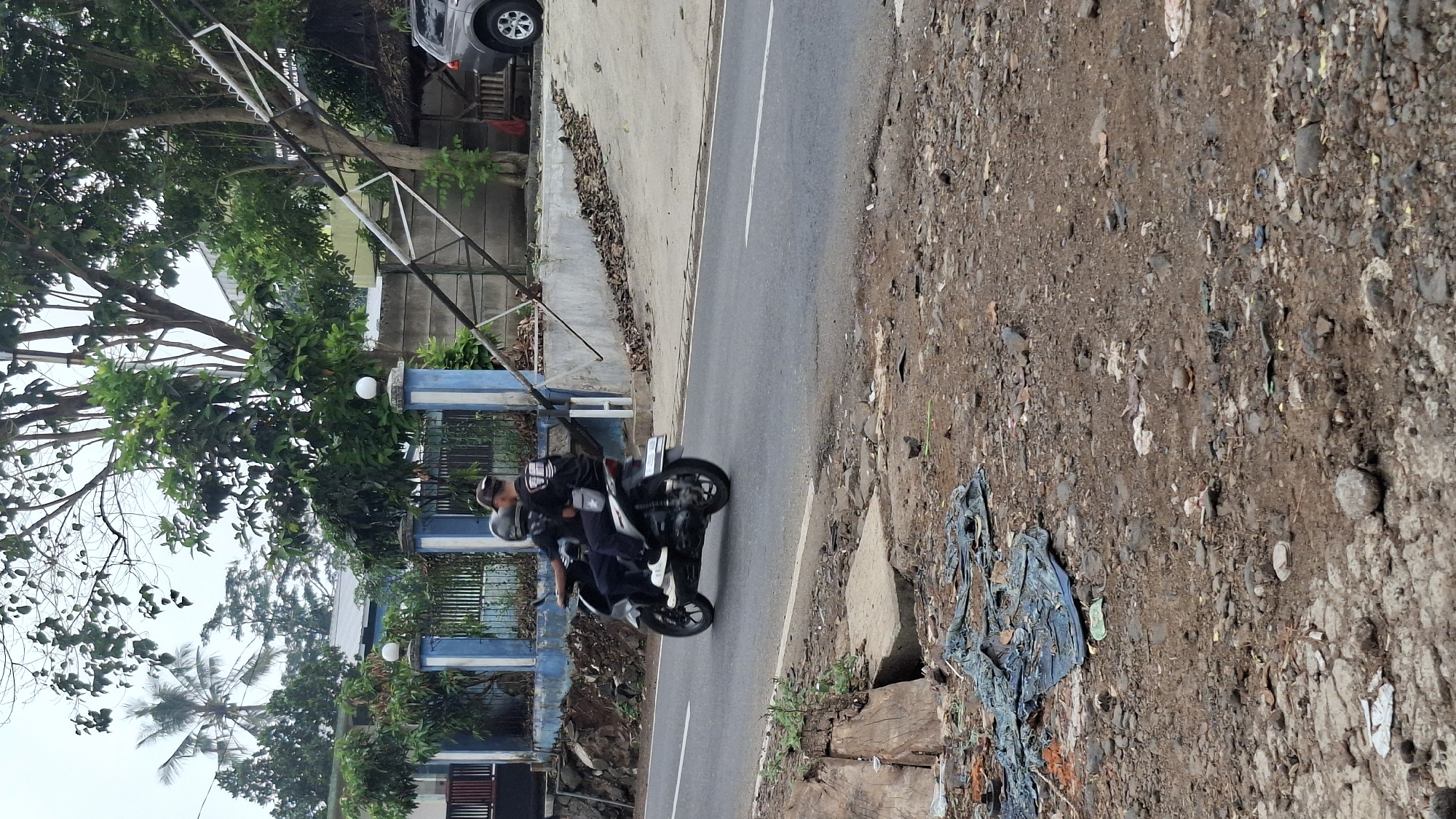 In Cilaku, Indonesia, two individuals are captured mid-journey on a motorcycle. The rider, wearing a black t-shirt with a graphic print and a baseball cap, steers the vehicle. The passenger, also in a black t-shirt and wearing a helmet, is seated behind the rider. They are traveling on a paved road, visible on the left side of the image, with a white line marking the lane.
The surrounding environment features a blue and white striped fence with decorative white globes atop pillars, suggesting a property boundary. Beyond the fence, lush green trees and foliage are abundant, with a tall palm tree visible in the background to the left. To the right, a grey SUV is parked. A damaged or fallen structure, possibly an electricity pole with diagonal white supports, is leaning precariously behind the fence. The foreground of the image depicts a rough, unpaved area littered with debris, including pieces of blue tarp, rocks, and other assorted rubbish. The sky is overcast, indicating a cloudy day.