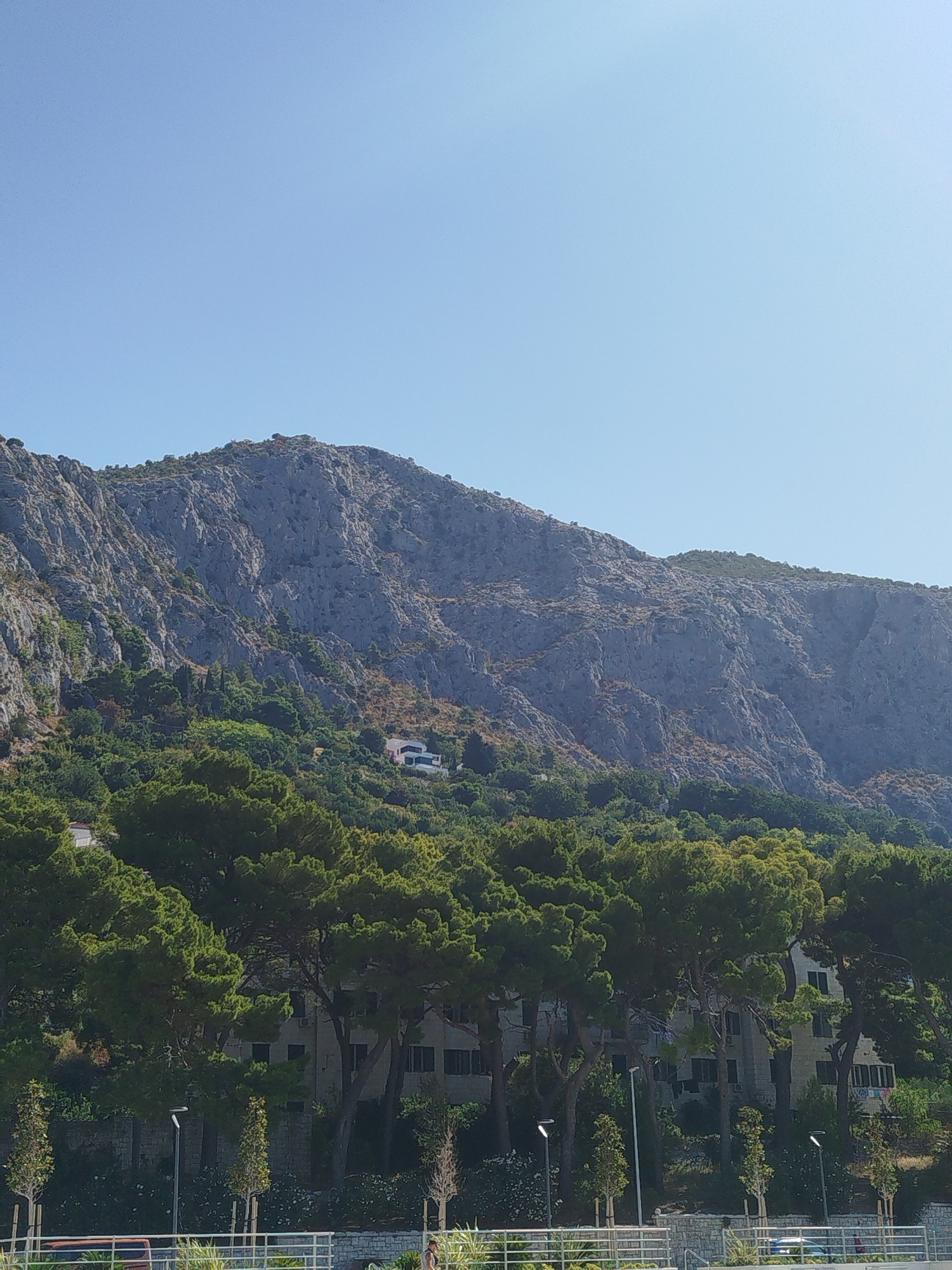 A bright, clear sky dominates the upper portion of the image, with a vast, rugged mountain range taking up the middle ground. The mountain's surface is textured with numerous rocky formations and sparse vegetation. Below the mountain, a dense forest of green trees creates a natural transition to the lower section of the image, which features a building complex partially obscured by the trees. The building appears to be several stories high with numerous windows. In the foreground, a white fence and some landscaping are visible, suggesting a public area or pathway.