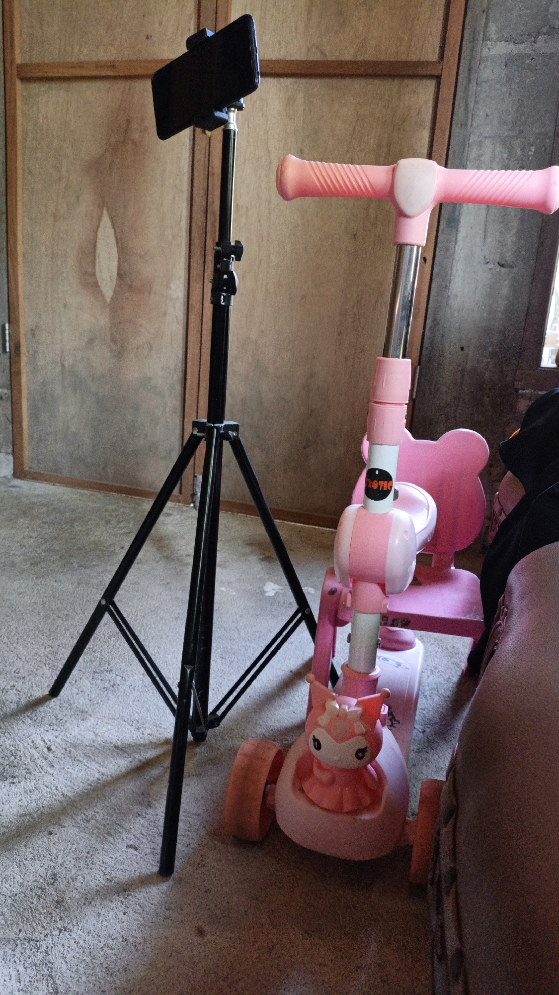 In Cikal, Indonesia, a pink children's scooter with a teddy bear-shaped backrest and a character figurine at the front is positioned next to a black tripod holding a smartphone. The scene is indoors, with a wooden door visible in the background and a concrete floor. The lighting suggests it is daytime, with shadows cast by the tripod and scooter.