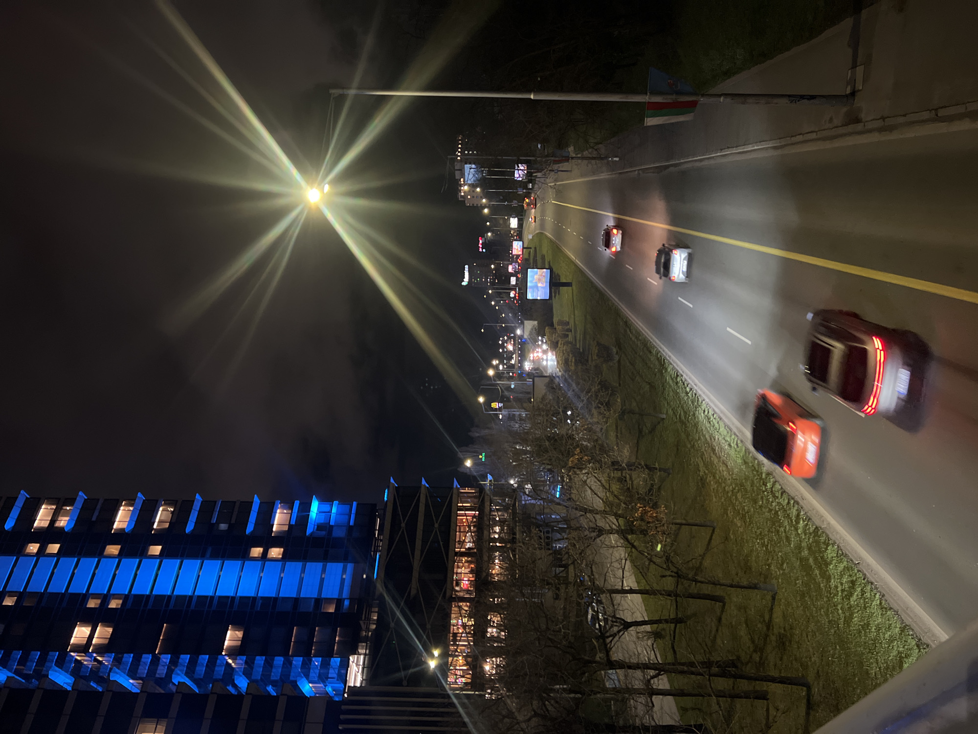 Here's a detailed description of the image:
**Overall Impression:** The image shows a night scene of a city road with cars in motion, captured from an elevated perspective. Buildings and streetlights illuminate the scene.
**Location:** Given the presence of the Azerbaijan flag, buildings and street design it is likely that this is an image of Heydar Aliyev Avenue in Sofia, Bulgaria.
**Specific Elements:**
* **Road and Traffic:** A multi-lane road is the primary element. Several cars are visible, with motion blur suggesting movement. A yellow line divides the traffic lanes.
* **Buildings:** On the left of the image, there's a tall building with blue illuminated sections. Further down the street, another building with brightly lit windows can be seen.
* **Streetlights:** Streetlights are positioned along the road, casting strong rays of light.
* **Landscaping:** A green strip of land runs alongside the road, with trees visible.
* **Other Details:** Billboards with advertisements are placed along the road. The flag of Azerbaijan is seen attached to a streetlight pole.
**Environment:** The scene takes place at night. The sky is dark and likely overcast.