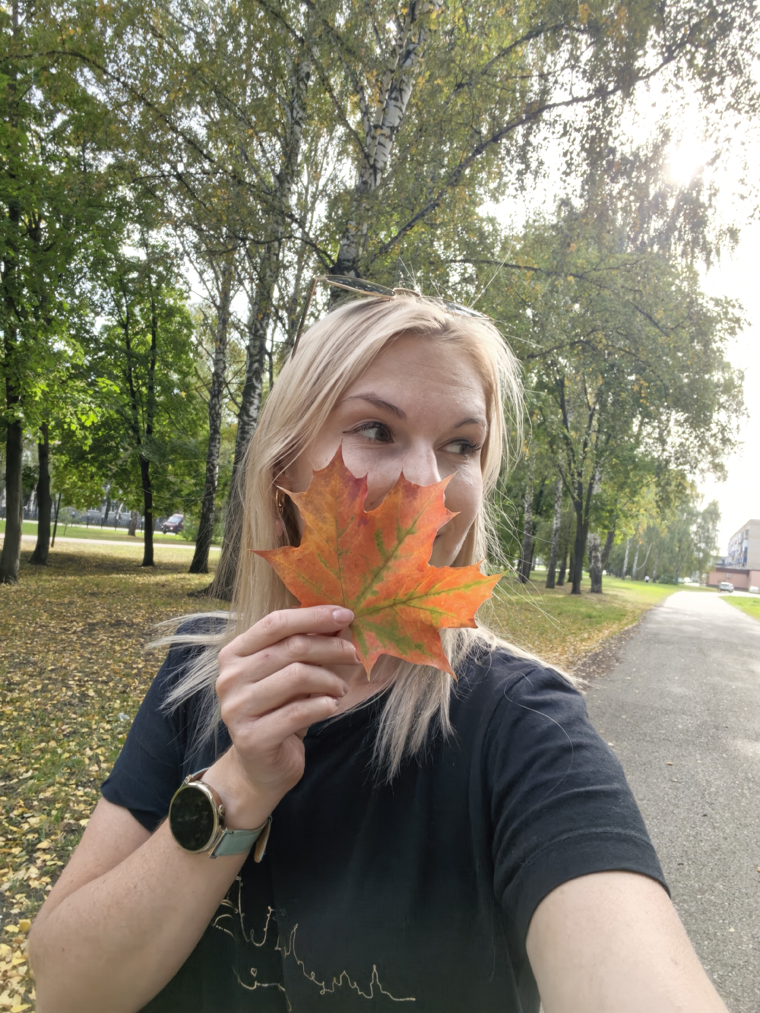 A blonde woman is holding a bright orange and green maple leaf in front of her face, obscuring part of her nose and cheek. She is looking to her right. She is wearing a black t-shirt and a smartwatch with a light blue band on her left wrist. The background is a park-like setting with trees, some with green leaves and others with yellowing leaves, and a paved path. The ground is covered with fallen yellow leaves. The sun is shining through the trees, creating lens flare. The scene appears to be outdoors in the city of Sterlitamak, Russia, during autumn.