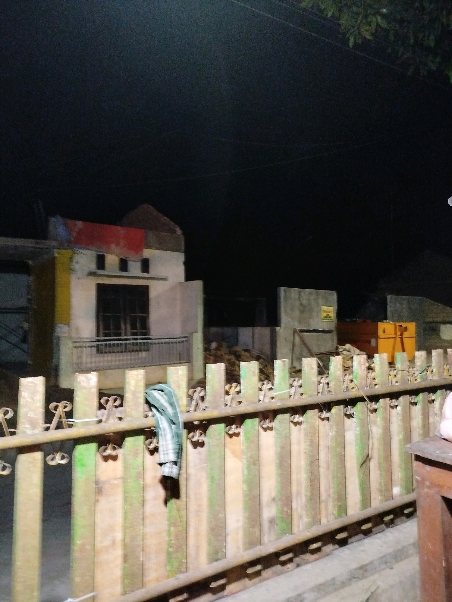 The image, taken at night in Mojopanggung, Indonesia, depicts a partially constructed or renovated building with a distinct red roof section. The building has a light-colored facade, with some areas painted yellow and other sections appearing unfinished. A dark-paned window is visible above a railing on what appears to be a balcony or porch. In the foreground, a rustic fence made of vertical wooden planks, some painted green, runs horizontally across the frame. A piece of checkered fabric, possibly clothing, is draped over the fence. Beyond the fence, construction materials and equipment, including what appear to be yellow and orange industrial containers or machinery, are visible in the background, along with concrete structures. The scene suggests ongoing construction or renovation activity at an outdoor location after dark.