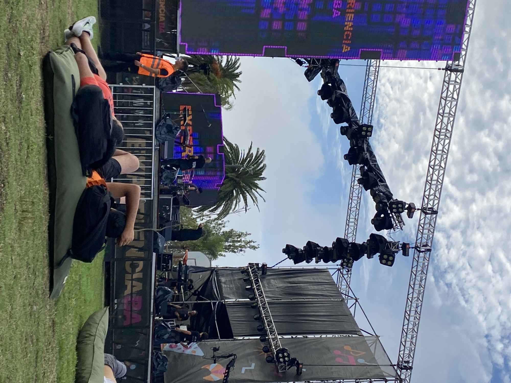 The image depicts an outdoor event in Buenos Aires, Argentina. A stage is visible with lighting rigs and speaker systems. A large LED screen displays text in Spanish. Palm trees are present. People are visible in the background, some wearing event staff apparel. In the foreground, an individual is reclining on a green cushion. The environment includes a grassy area.