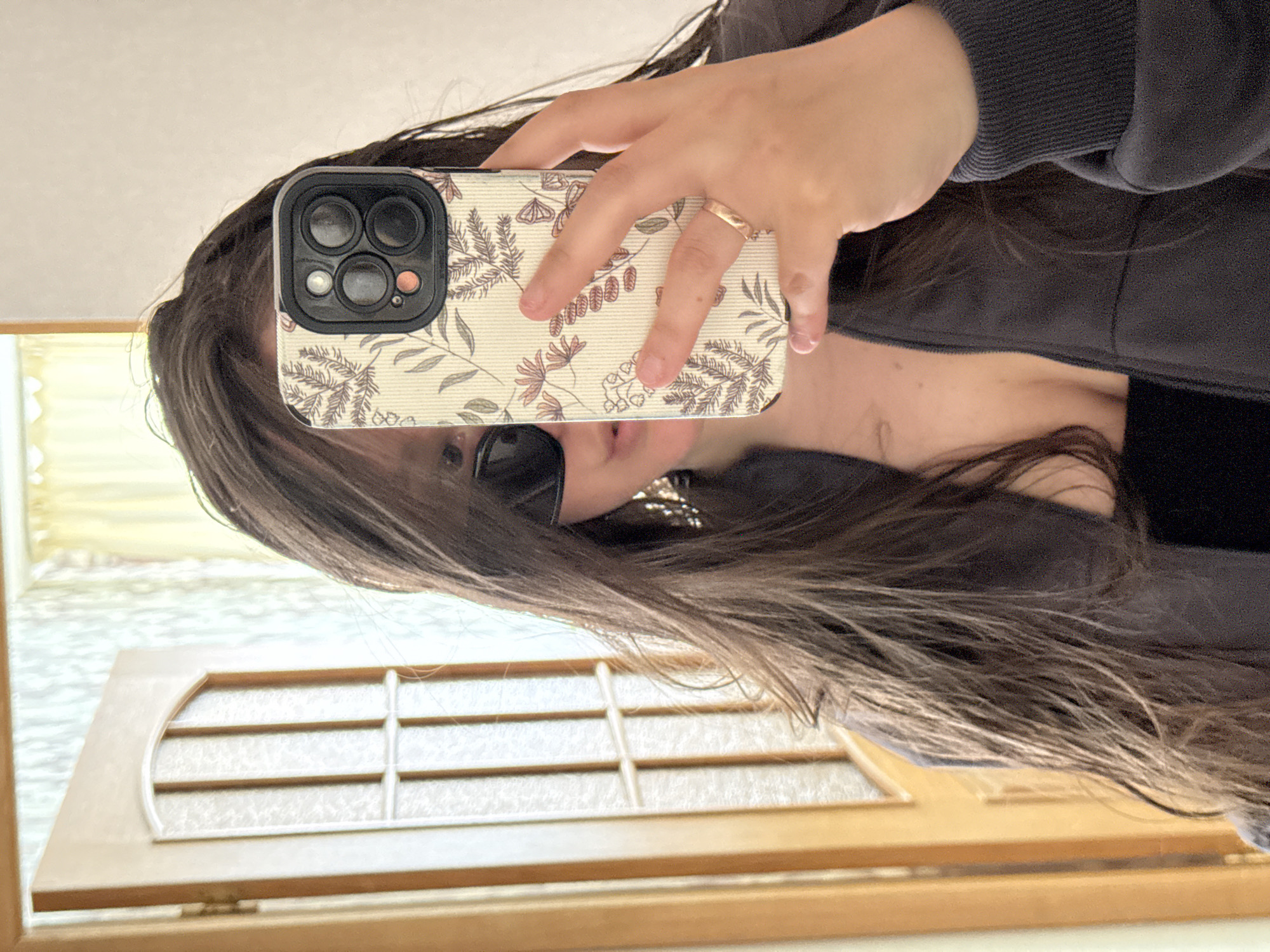 A person wearing a grey top and sunglasses is taking a selfie in a mirror with a smartphone in a floral-patterned case. The reflection shows a door with small square panes. The location is undetermined and there is no indication this photo was taken in Krasnodar, Russia.