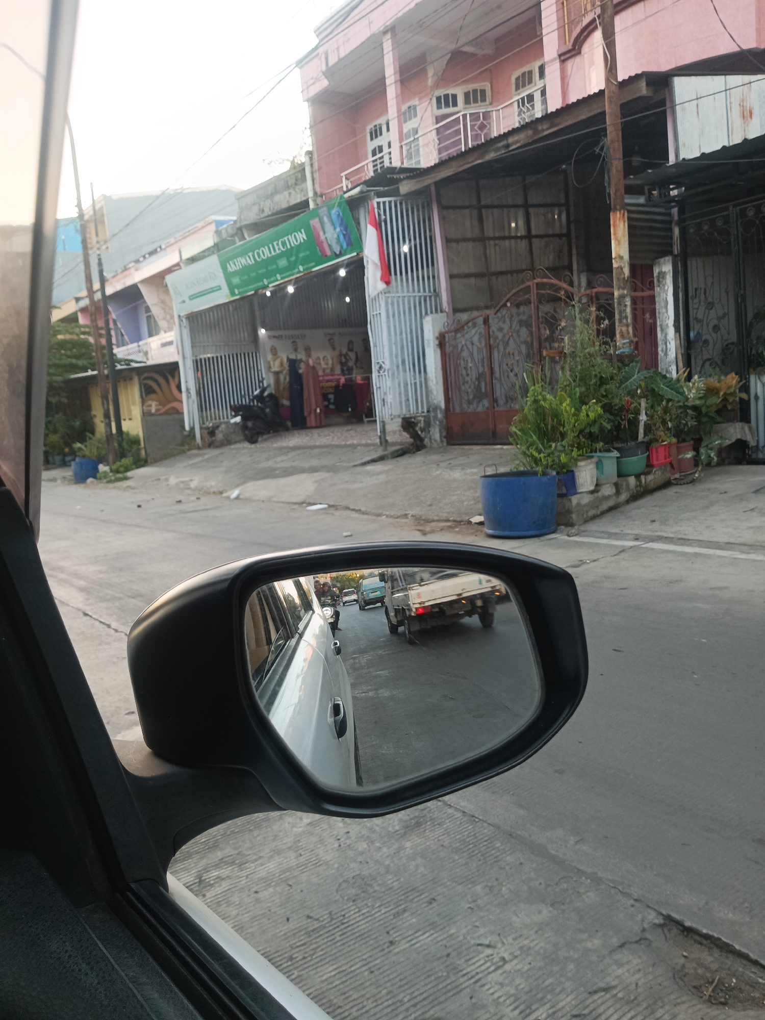 This is a medium-shot, eye-level, exterior, daytime photograph taken from inside a vehicle, looking out a side window towards a rearview mirror reflecting a street scene. The photograph is set in Pangkajene, Indonesia, indicated by visual cues such as the architecture and the presence of Indonesian flags.
In the foreground, the black housing of a car's side rearview mirror is prominent, angled towards the left of the frame. The mirror reflects a street with other vehicles. A white car, presumably the one the photo is taken from, is partially visible to the left of the mirror.
The background shows a street with a slight incline. On the right side of the street, there are buildings with pink and grey facades. One building has an overhang supported by a wooden pole. In front of the buildings, there is a row of potted plants, including large green foliage in blue, white, and green pots. A large blue plastic container is also visible near the plants. Further back, a metal gate is visible.
On the left side of the street, there are other buildings, some with balconies and windows. A green banner hangs above a storefront, reading "AKIHWAT COLLECTION" in white lettering. Below the banner, there are displays of clothing and mannequins. A black motorcycle is parked in front of the storefront. A small Indonesian flag is attached to the building. The sky is bright and appears to be overcast or hazy.
The overall scene depicts a typical street in a residential or commercial area of Pangkajene, Indonesia, with a focus on the view through a car window and its reflection.