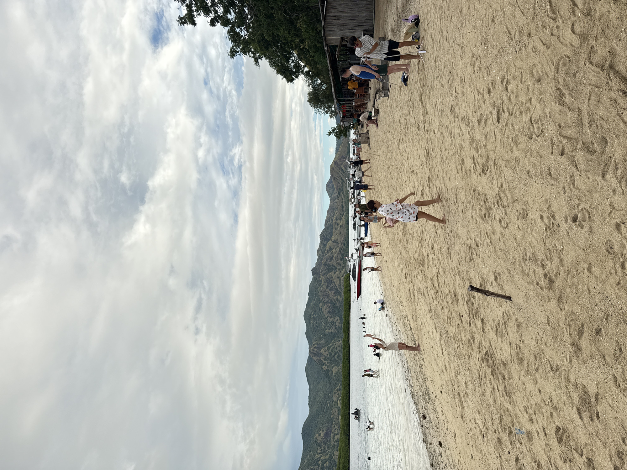 The image depicts a beach scene in Rinca Village, Indonesia. The foreground features a sandy beach with multiple footprints and a piece of driftwood. People are scattered across the scene, some wading in the water and others standing on the sand. In the midground, several boats are docked near the shore. In the background, there are green mountains under a cloudy sky. At the top left corner of the image, there are trees and a building.
