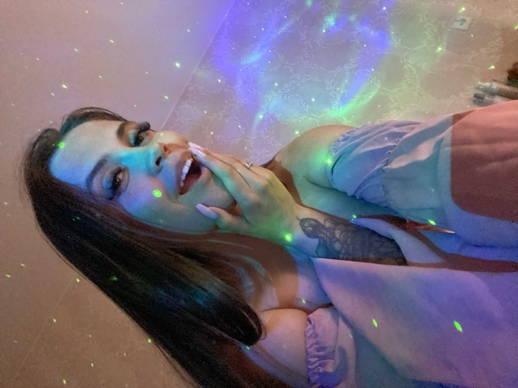 An over-the-shoulder, medium shot captures a young woman with long, dark hair, smiling with her mouth open and hand touching her cheek. She is wearing a light purple, off-the-shoulder top with ruffled sleeves. The background is a textured wall with a faint floral pattern, illuminated by a light projector creating green and purple star-like effects. The lighting is dim and atmospheric. There are no visible indications of the city of Chaikovsky, Russia.