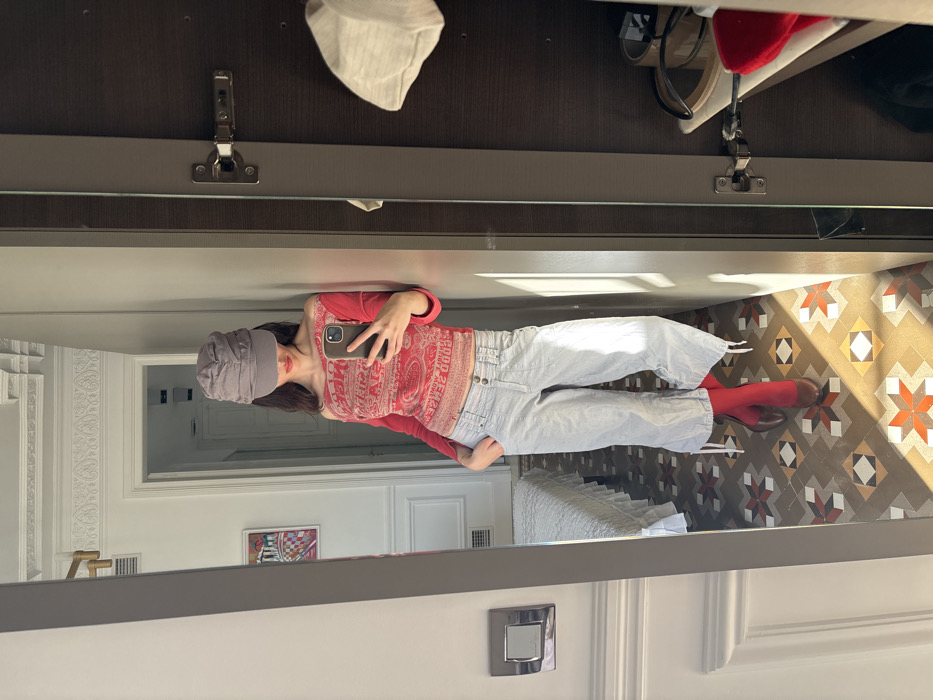 A female individual is depicted taking a mirror selfie in an indoor setting. She is wearing a grey newsboy-style cap, a long-sleeved red off-the-shoulder top with a lighter faded pattern, light-colored cropped trousers with ties at the hem, red socks or hosiery, and brown high-heeled boots. Her left hand is partially inserted into her trouser pocket. Her right hand holds a silver-colored smartphone, pointed towards the mirror to capture the image.
The environment is a room characterized by white walls featuring decorative wainscoting and ornate crown molding. The floor is covered with distinctive patterned hydraulic tiles (baldosa hidráulica), displaying geometric shapes in shades of brown, beige, orange, and red. This flooring is characteristic of architecture in Barcelona, Spain. Natural light illuminates the room, casting strong shadows on the floor. Reflected in the mirror, a doorway leads to an adjacent room where a bed or bench with a white ruffled cover and a framed abstract artwork on the wall are visible. A silver-colored wall switch or outlet is affixed to the wall on the left side of the reflection. To the right of the mirror, a dark brown wardrobe door is partially ajar, revealing a shelf containing various items, including a red fabric object, and a light-colored cloth hanging on the inner door panel. A metal hinge is visible on the wardrobe door.