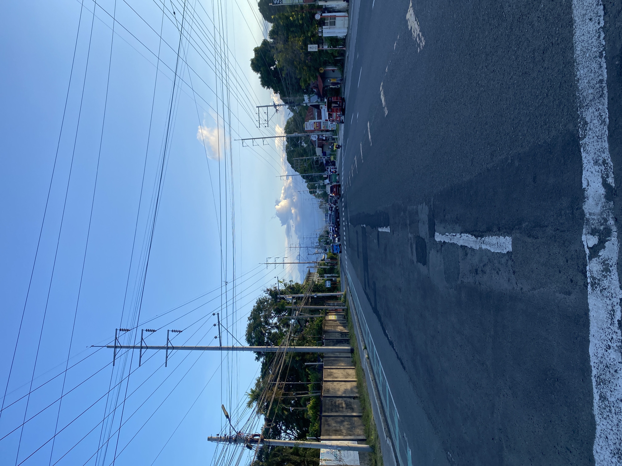 The image captures a wide-angle view of a busy street in Davao City, Philippines, likely during the late afternoon or early evening, given the quality of light. The road appears to be a multi-lane highway, with vehicles including cars and a red jeepney visible in the distance. Overhead, a complex network of utility poles and wires crisscrosses the sky, a common sight in many Philippine cities.
On the left side of the road, a concrete wall runs alongside a paved walkway, possibly a pedestrian path or a bike lane marked with green and white lines. Beyond the wall, lush green trees and vegetation are visible, suggesting a suburban or semi-urban setting.
On the right side of the road, commercial establishments with various signage are present, indicating a developed area. A few individuals can be seen near these establishments, and a tricycle-like vehicle is also present, typical of public transportation in the Philippines.
The sky is a clear blue, with a few scattered clouds in the distance, suggesting fair weather. The overall impression is a snapshot of daily life and infrastructure in a dynamic urban environment.