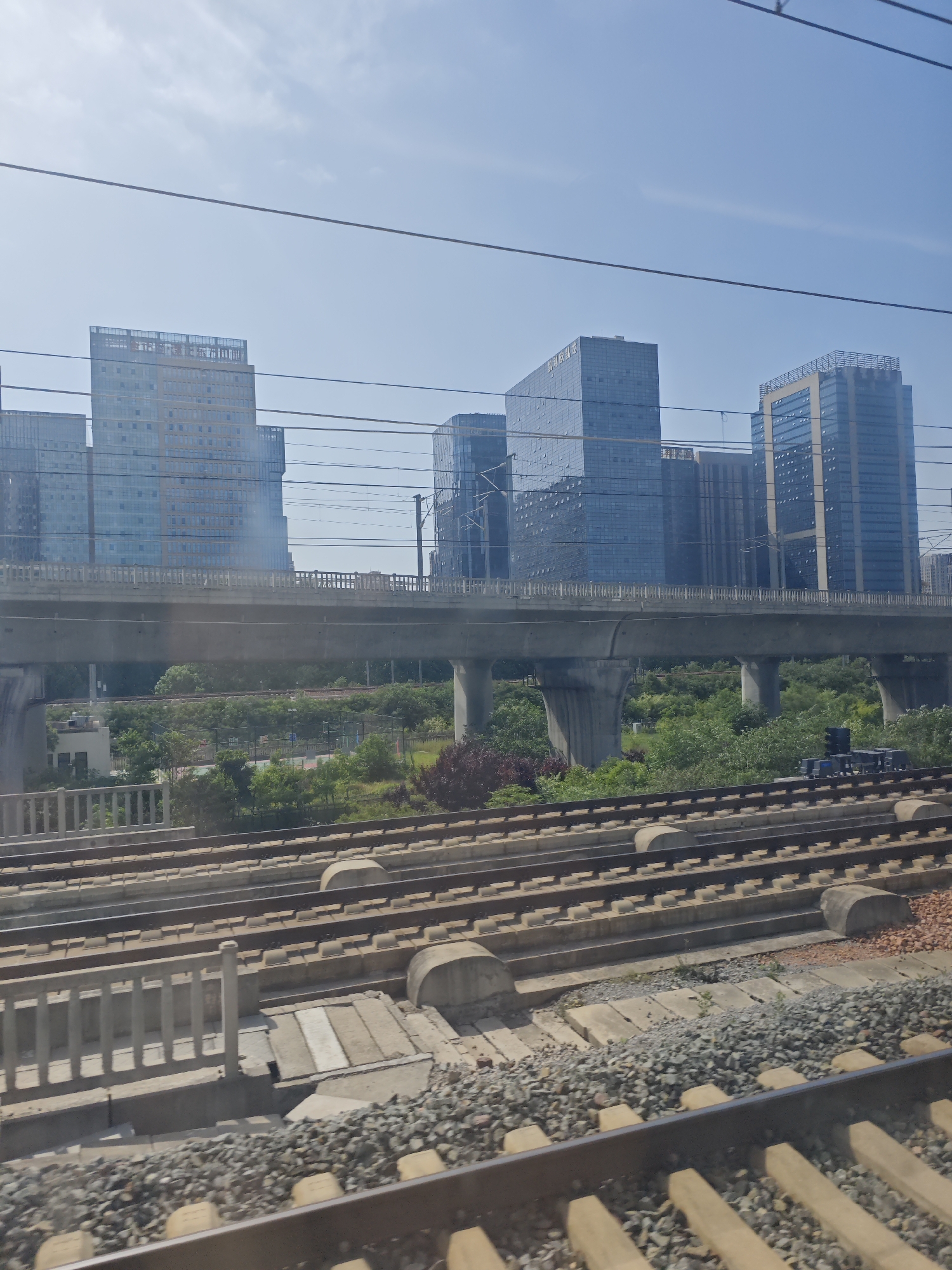 The image presents a modern urban landscape, likely captured from a vantage point near or on a train. In the foreground, multiple parallel sets of train tracks, laid on concrete sleepers and gravel ballast, stretch across the bottom of the frame. A low concrete barrier or railing is visible beside some of the tracks.
The midground is dominated by a substantial elevated concrete overpass, supported by thick pillars, which spans horizontally across the scene. This overpass carries what appears to be another set of railway tracks or a multi-lane road. Beneath the overpass and in the spaces between the various transportation layers, dense green vegetation, including trees and shrubs, thrives, interspersed with smaller concrete structures.
The background showcases a skyline comprised of numerous modern high-rise buildings, most featuring reflective blue or grey glass facades and contemporary architectural designs. These buildings suggest a commercial or office district. On one of the prominent glass buildings, faint Chinese characters "中国移动" (China Mobile) are discernible.
The upper portion of the image reveals a bright, clear blue sky with only a few faint clouds, traversed by thin utility wires. The scene depicts a sunny daytime environment, highlighting the layered infrastructure and modern development typical of a large city. No people are visible.