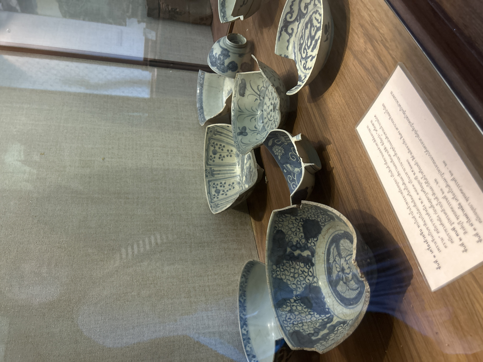 A close-up shot features several broken blue and white porcelain bowls displayed on a wooden surface behind glass. The bowls exhibit intricate patterns and appear to be ancient artifacts. A white placard with Thai text is visible in the foreground, partially obscuring some of the bowls. The background consists of a textured, light-colored fabric. The lighting suggests an indoor setting, likely a museum or exhibition.