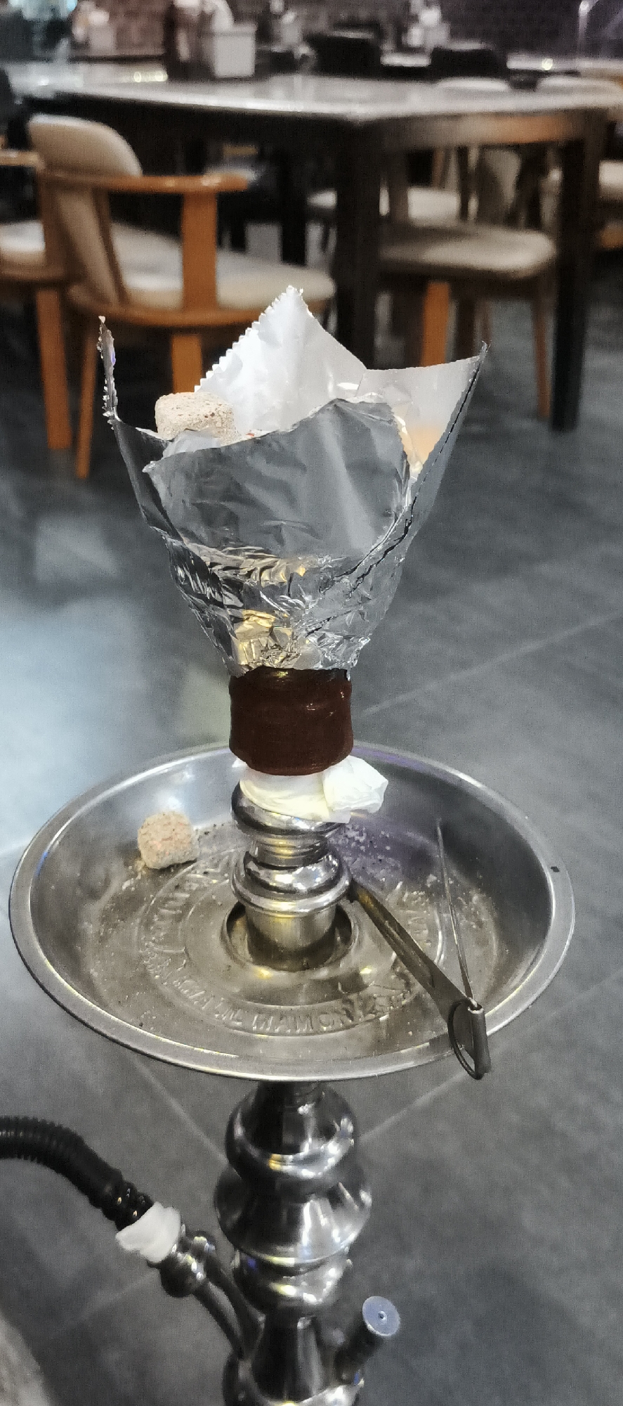 The image captures a close-up of a hookah (shisha) setup in what appears to be an indoor cafe or lounge in Bangkok, Thailand. The primary subject is the upper section of a silver-toned hookah.
At the top, a dark brown, possibly clay or ceramic, bowl is covered with crinkled aluminum foil. Several white, cube-shaped coals are placed on the foil, indicating that the hookah is either in active use or has just been prepared. Below the bowl, a circular, metallic ash tray shows signs of use, with scattered ash or residue. A pair of silver tongs, used for handling coals, rests on the tray, alongside a single unlit or spent coal cube. A small piece of white tissue or paper is visible between the bowl and the main stem, possibly for stability or to catch drips.
The hookah's stem, made of polished silver metal, reflects the surrounding dim light. A black hose is connected to the lower part of the stem on the left, with a white, disposable mouthpiece visible at the connection point.
The background is softly blurred, revealing a dining or lounge area with dark wooden tables and chairs. Some chairs have light-colored seat cushions and wooden frames, some light brown and some dark. The dim lighting suggests an evening or nighttime setting. The floor appears dark and possibly tiled. Although no people are distinctly visible, the arrangement implies a social gathering. Partially visible text on the ash tray reads "...L DIAMON...", possibly part of a brand name like "Khalil Mamoon".