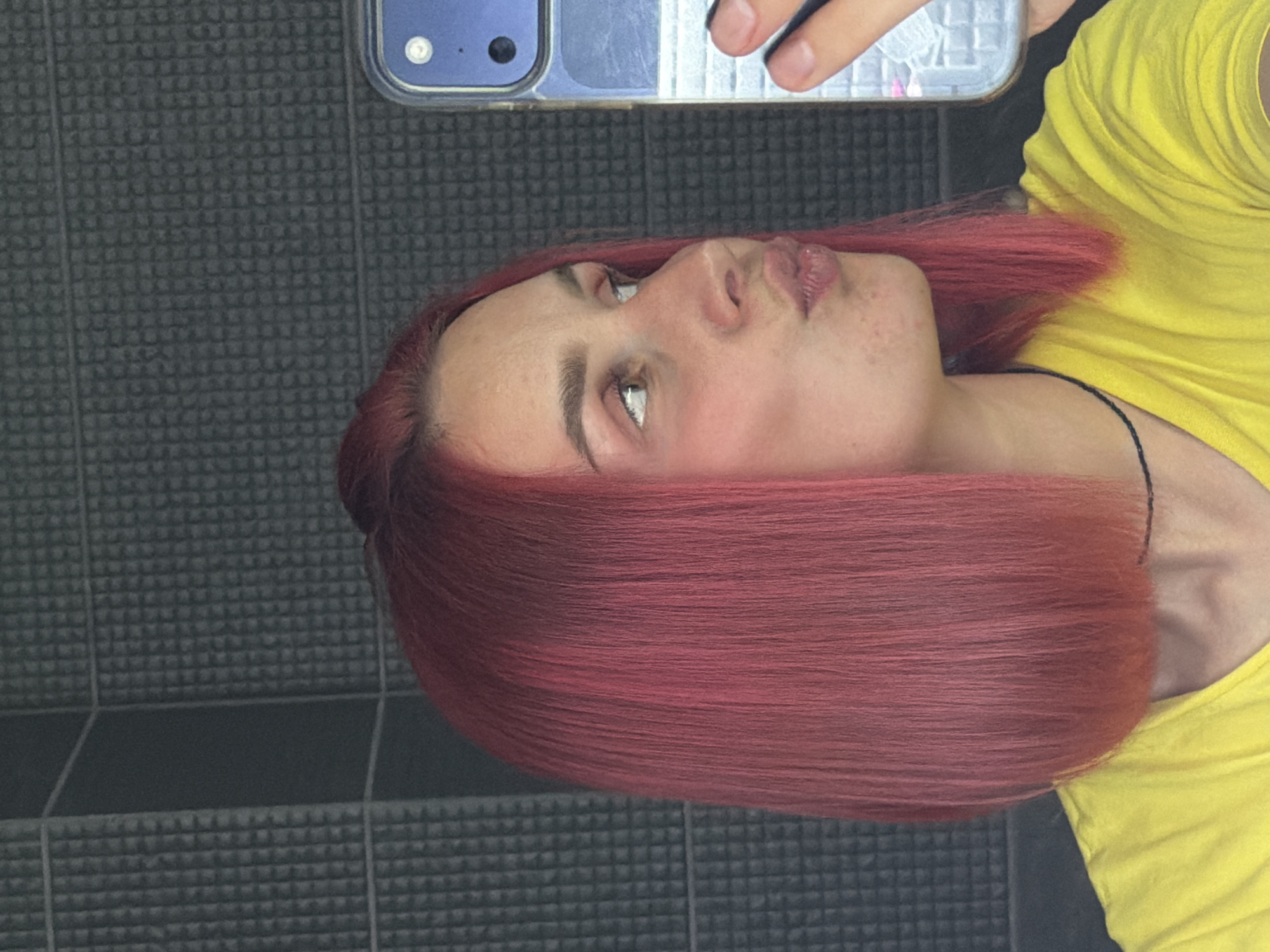 The image shows a young woman with pink hair taking a selfie in a bathroom. The wall behind her is tiled with dark gray square tiles. She is wearing a yellow t-shirt and a thin black necklace. Her lips are pursed, and she is looking at her phone, which is visible in her right hand. The bathroom appears to be in an apartment or house. There is no information in the image to suggest the location is in Chaikovsky, Russia.