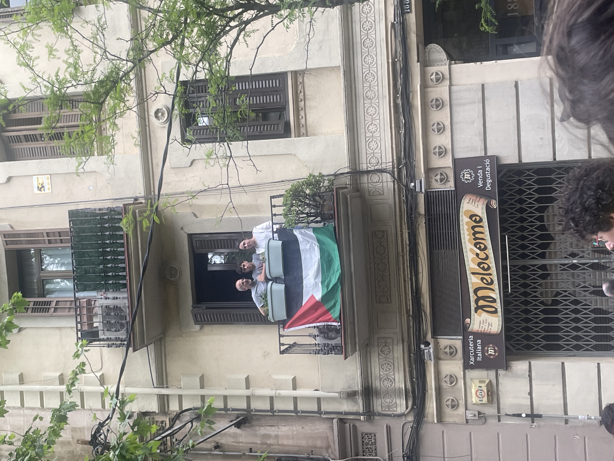 The image captures a daytime scene of a multi-story building facade in what appears to be an urban setting in Barcelona, Spain. Lush green tree branches partially frame the view from the top and left.
On a second-story balcony, three men are visible, casually dressed and looking downwards. A large Palestinian flag is prominently draped over the balcony railing, displaying its black, white, green, and red colors. Potted plants also adorn the balcony. The men's expressions are neutral, suggesting they are observing something below or simply standing.
Below the balcony, at street level, is a commercial establishment. A large brown sign with yellow lettering reads "Melocom." Below this, the text "Venda i Degustació" (Sale and Tasting) and "Xarcuteria Italiana" (Italian Charcuterie) indicates the shop sells Italian deli products. The shopfront is secured by a metal grate.
The building itself features classic architectural details, including decorative moldings, patterned elements, and traditional wooden window shutters. Various electrical wires run across the facade. A small white plaque with the number "100" is visible to the left of the balcony, and the number "188" is carved into the building's upper right section. The overall lighting suggests it is daytime, possibly under an overcast sky. Part of a person's head and hair is visible in the bottom right foreground, indicating the photo was taken from a crowded or close perspective.