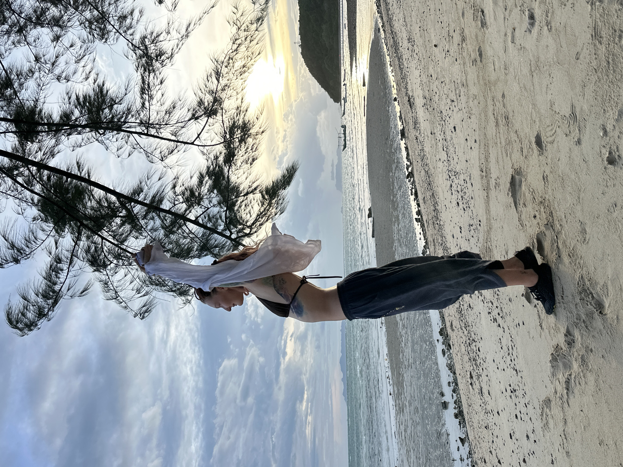A medium shot captures a woman standing on a sandy beach in Ko Samui District, Thailand, during what appears to be sunset or sunrise. She is facing left, her body angled slightly away from the camera. She wears a dark bikini top and dark, loose-fitting pants. Her arms are raised, holding a sheer white garment that is billowing in the wind. Her hair is reddish-brown and pulled back. The background shows the ocean with gentle waves, a distant shoreline with a hill, and several boats. The sky is cloudy with the sun low on the horizon, casting a soft light. A tree with thin branches hangs into the frame from the top right, its needles also appearing to move in the wind. The sand beneath her feet shows some texture and small debris.