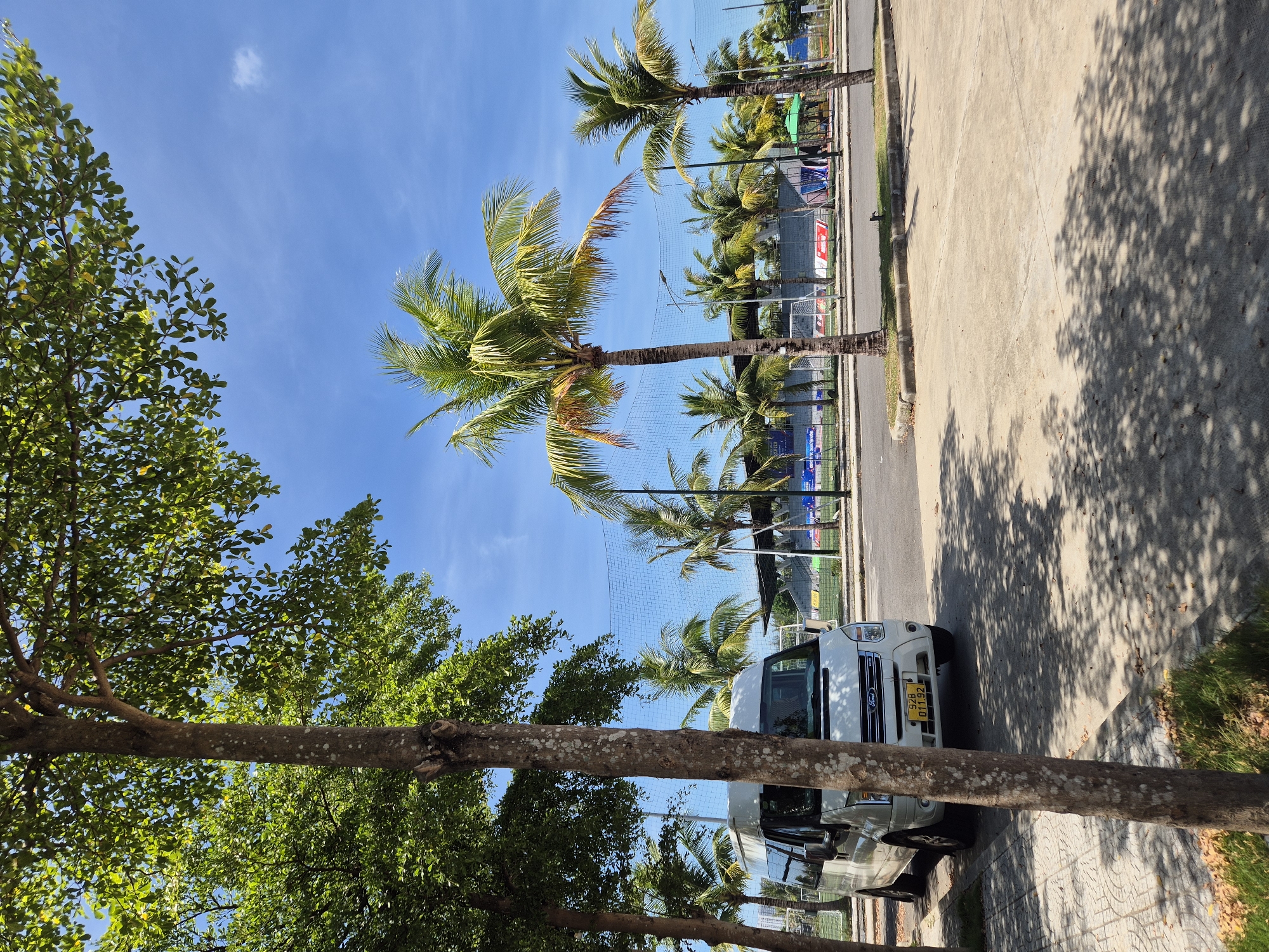 A white Ford Transit van is parked on a concrete surface next to a paved road, with a sidewalk to the left. The van has a yellow license plate reading "928 011.92". In the background, several palm trees are visible, along with a fenced sports field that appears to be a soccer field, evidenced by a goal post. There are bleachers and a covered seating area in the distance. The scene is under a bright blue sky with scattered white clouds, and dappled sunlight casts shadows from trees onto the ground. The location is the beachside area in Đà Nẵng, Vietnam, likely near the East Sea Park or along the coast road.
