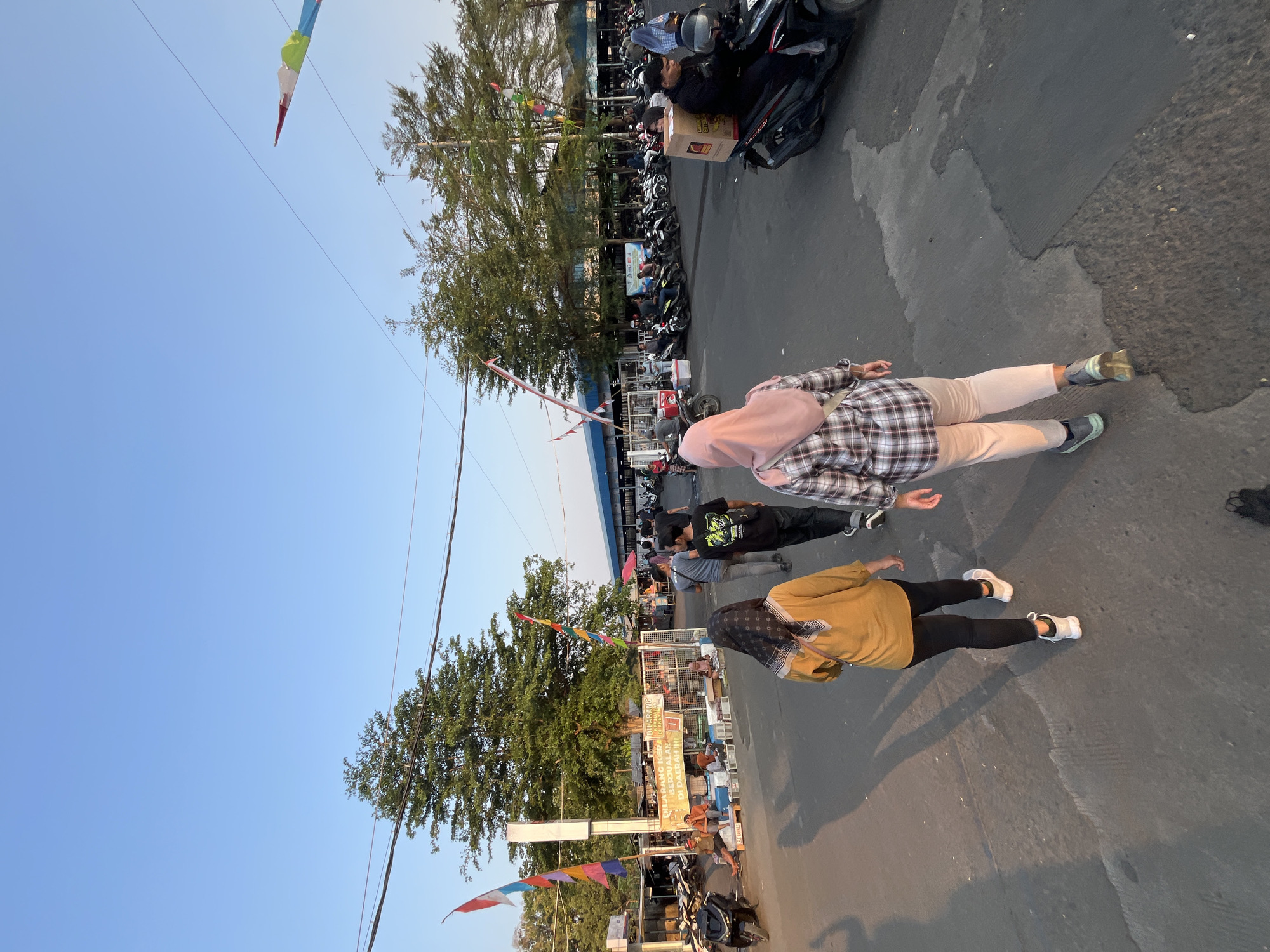 The image depicts a street scene in Pelang, Indonesia. The weather is clear with a blue sky. In the foreground, two women are walking away from the camera. The woman on the left is wearing a yellow long-sleeved shirt, black pants, and white sneakers, with a patterned scarf on her head. The woman on the right is wearing a plaid long-sleeved shirt, light-colored pants, and sneakers, with a pink hijab. Behind them, several other people are visible, some walking and some sitting.
The street is paved with asphalt and shows signs of wear and tear. On the left side of the street, there are market stalls with various goods, including some wrapped in plastic. Several motorcycles are parked or moving in the area. Flags, including Indonesian flags and colorful pennants, are visible in the background. Trees line the street, and a building with a blue roof is seen in the distance. The lighting suggests it is late afternoon or early evening, with long shadows cast on the ground. One person is riding a motorcycle with a large cardboard box strapped to the back. The overall impression is that of a bustling local market or street.