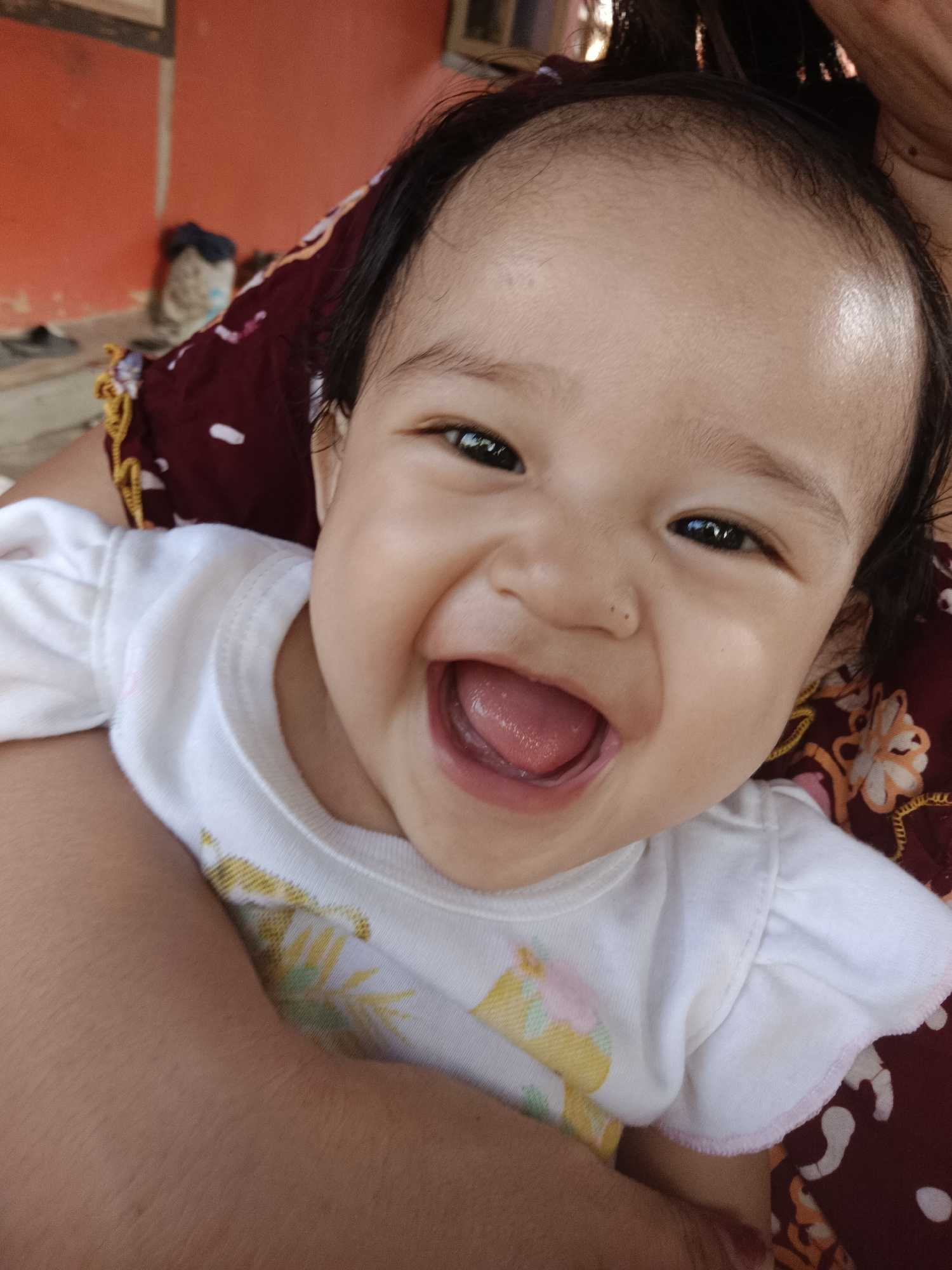 The image is a close-up of a baby's face, who is smiling with their mouth open, showing their tongue. The baby has dark hair and dark eyes. They are wearing a white shirt with a floral pattern. The baby appears to be held by someone, as parts of an arm and a patterned cloth are visible in the foreground and background. The background is blurry but suggests an indoor or outdoor setting with an orange wall and some objects that are not clearly identifiable. The image is taken from a high angle, looking down at the baby. Based on the provided information, the location is Kawua, Indonesia.