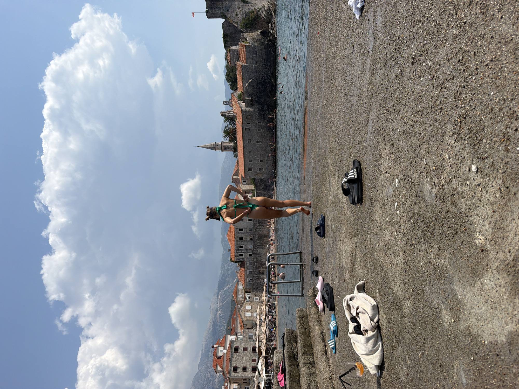 A woman in a green bikini stands on a stone pier in Budva, Montenegro, with her back to the camera. She is adjusting the back of her bikini bottoms. In the background, the Adriatic Sea is visible, with people swimming. Beyond the water, the historic Old Town of Budva is seen, featuring stone buildings with red-tiled roofs, a church steeple, and a fortress. Mountains rise in the distance. Scattered on the pier are various flip-flops and a towel with a glass of orange liquid and a straw. The sky is blue with scattered white clouds.