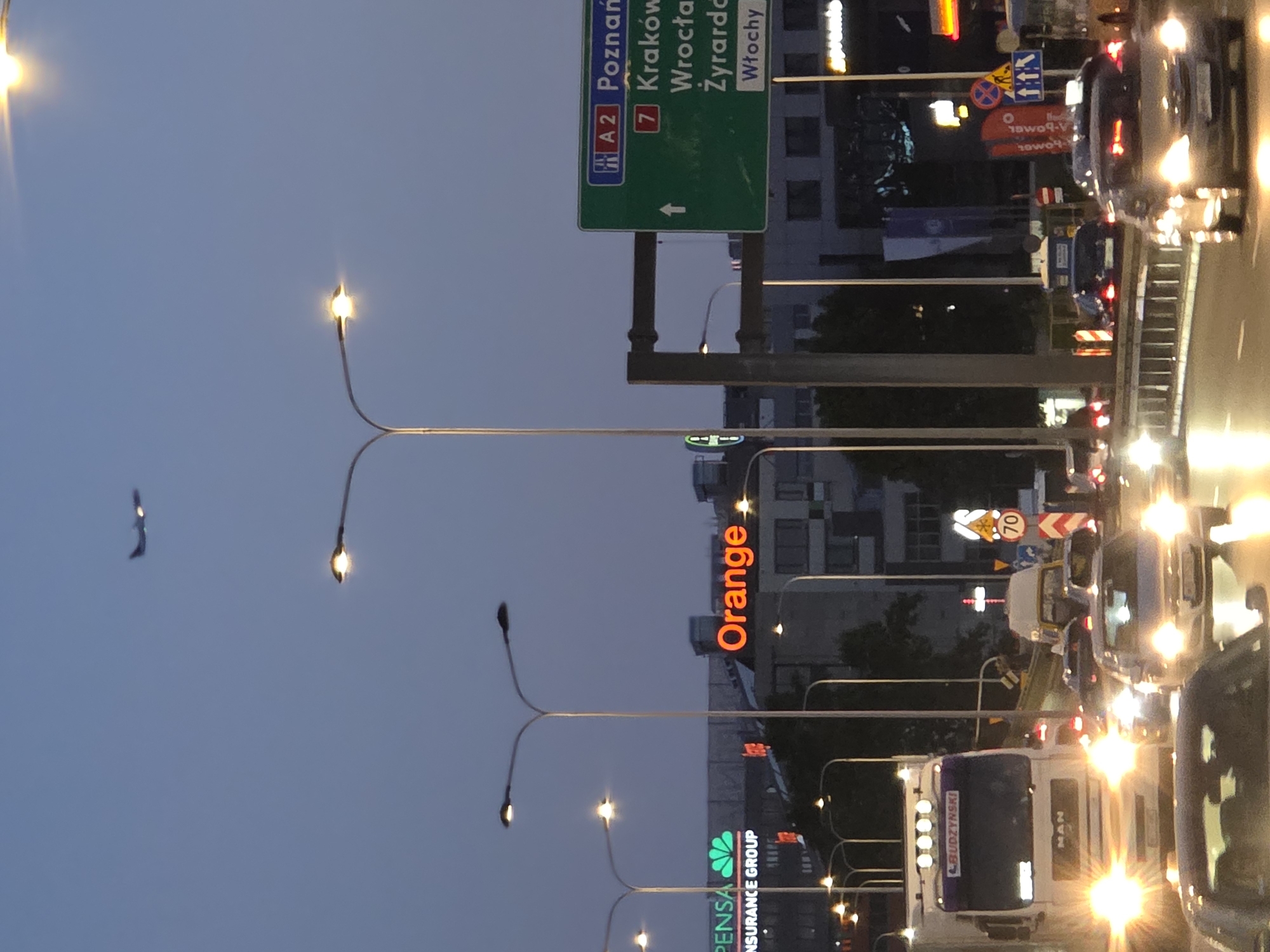 The image depicts a city street scene at dusk or night, likely in Poland, given the visible text on the road signs. The perspective is from within a vehicle, looking forward. The scene is dominated by the glow of streetlights, vehicle headlights, and illuminated signs, creating a vibrant and slightly blurred effect due to motion or low light.
Several objects are prominent:
- **Road Signs:** A large green road sign indicates directions to "Poznań," "Kraków," "Wrocław," "Żyrardów," and "Włochy," with the road number "A2" and "7" also visible. Another sign shows a red circle with a "70" inside, indicating a speed limit.
- **Vehicles:** Numerous cars and a white truck are visible, caught in traffic. The truck has "BUDZYNSKI" written on its front and "MAN" on its side. Headlights and taillights of various vehicles are bright and reflective.
- **Buildings:** A multi-story building with illuminated windows and a prominent orange sign that reads "Orange" is visible in the background. Another building below it has a sign that says "PENSA INSURANCE GROUP."
- **Streetlights:** Multiple streetlights with multiple lamps extend over the road, some with visible lights and others appearing as silhouettes against the sky.
The overall atmosphere is that of a busy urban environment at night, with traffic illuminated by artificial lights. The time of day is clearly after sunset, with the sky displaying a deep twilight blue. There is no indication of weather conditions, but the lighting suggests it is a clear evening. The focus is on the transportation infrastructure and commercial signage of a city.