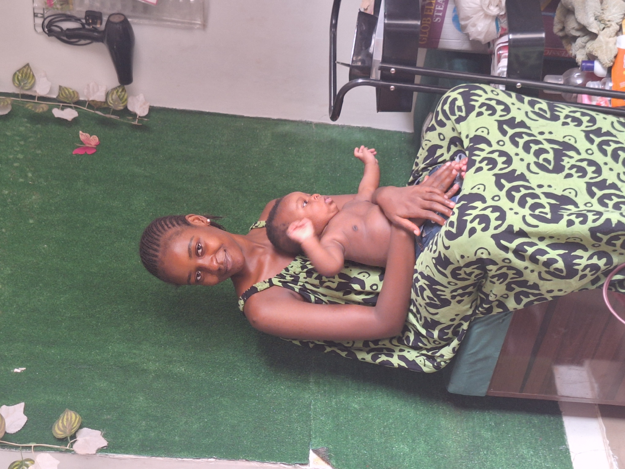 An indoor, eye-level shot captures a young woman and a baby in what appears to be a relaxed and affectionate moment. The scene is likely set in a home or establishment in Sabon Tasha, Nigeria.
The main subjects are a young woman, possibly in her late teens or early twenties, and an infant. The woman is lying down, supporting herself on her elbow, and smiling directly at the camera. Her hair is styled in neat braids, and she wears a light green dress or top with a distinctive dark green or black abstract pattern. She is holding the baby on her chest/stomach area. The baby, appearing to be a few months old, is bare-chested and looking upwards and slightly to the right of the camera, away from the viewer. The woman's expression is warm and happy, conveying a sense of tender connection with the infant.
They are resting on a vibrant green surface that resembles artificial turf or a mat, which covers the floor. In the background, along the wall behind them, decorative elements are visible: a white and green leafy vine with a small pink butterfly adorns the wall. To the left, a black hairdryer is mounted on the wall. To the right, a dark metal rack or shelf holds various items, including a white plastic bag, some folded fabric, and several bottles of different sizes and colors. Partial text reading "GLOB EL..." and "STEAL..." is visible on an item within the rack. A dark wooden surface, possibly a table or cabinet, is also seen in the lower right portion of the frame.
The lighting is soft and diffused, suggesting an indoor setting with either natural light filtering in or ambient artificial light. The overall mood is one of comfort and joy.