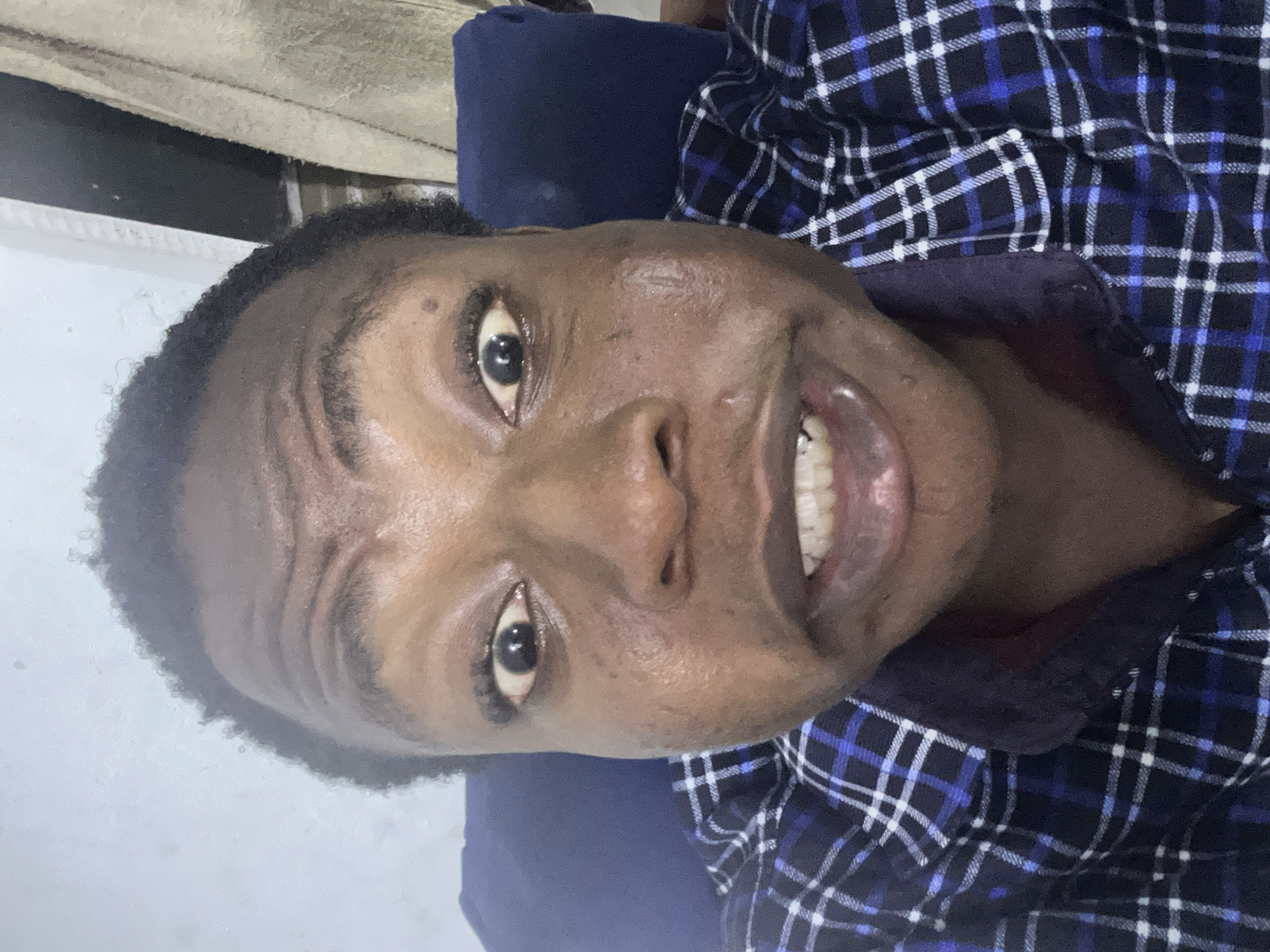 A close-up portrait of a Black person, possibly male, is presented in a tilted orientation. The individual is smiling, exposing the teeth. The person is wearing a dark blue and white plaid shirt. The background appears blurred and consists of neutral tones, potentially a wall or bedding. The location is not discernible.
