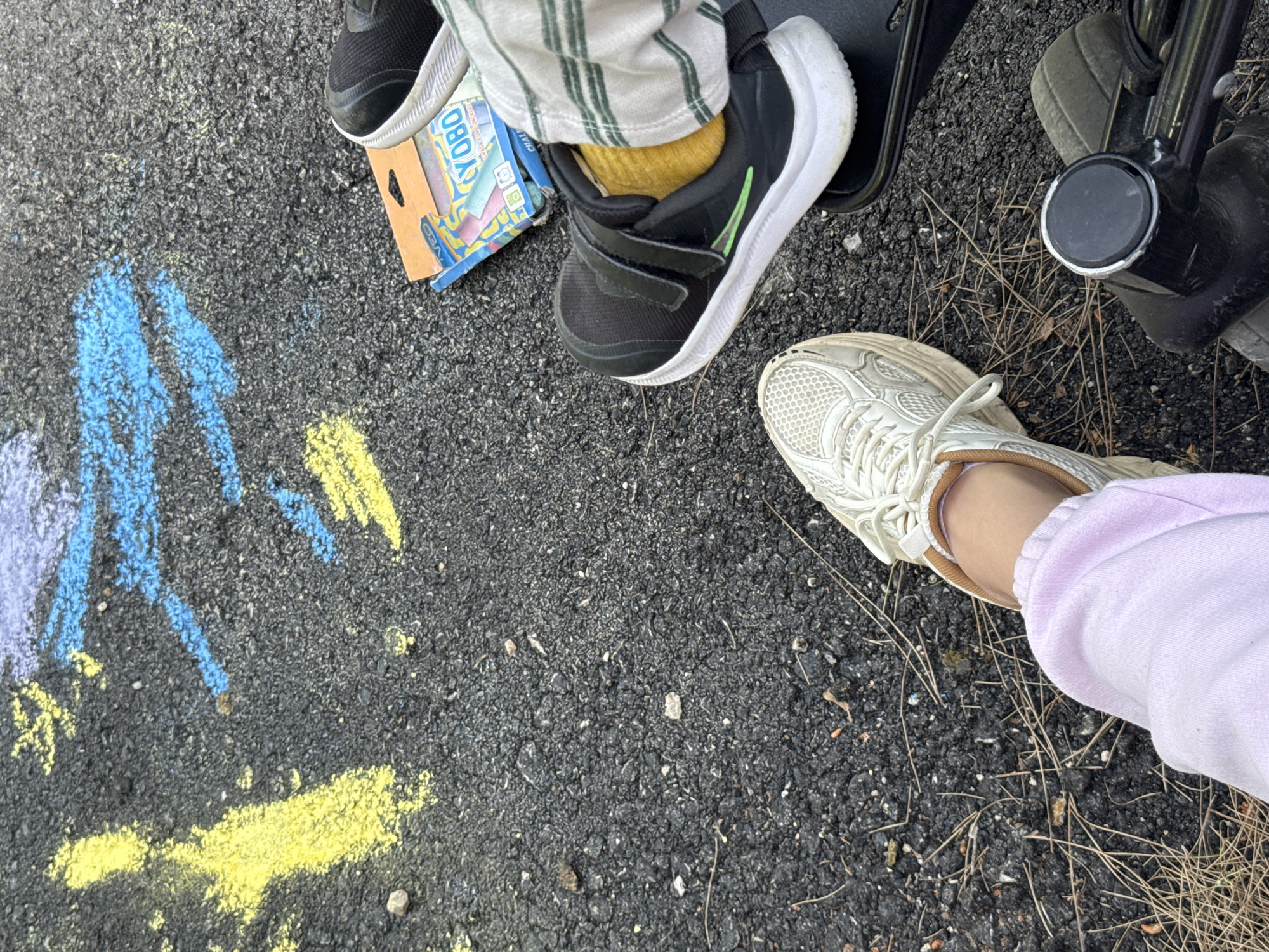 The scene is outdoors on a black asphalt surface, possibly a sidewalk or parking lot. There are remnants of blue and yellow chalk drawings on the left side of the frame. Scattered dry grass and debris are present on the asphalt.
The lower right corner shows a person's foot in a white sneaker and light purple sweatpants. Adjacent to this is a leg and foot wearing a black sneaker with a green stripe and a yellow sock. The leg is covered in striped pants. A box of "YOBO" chalk sits next to the sneakers. To the right of the chalk is a black sandal and a part of a black walking aid or stroller.