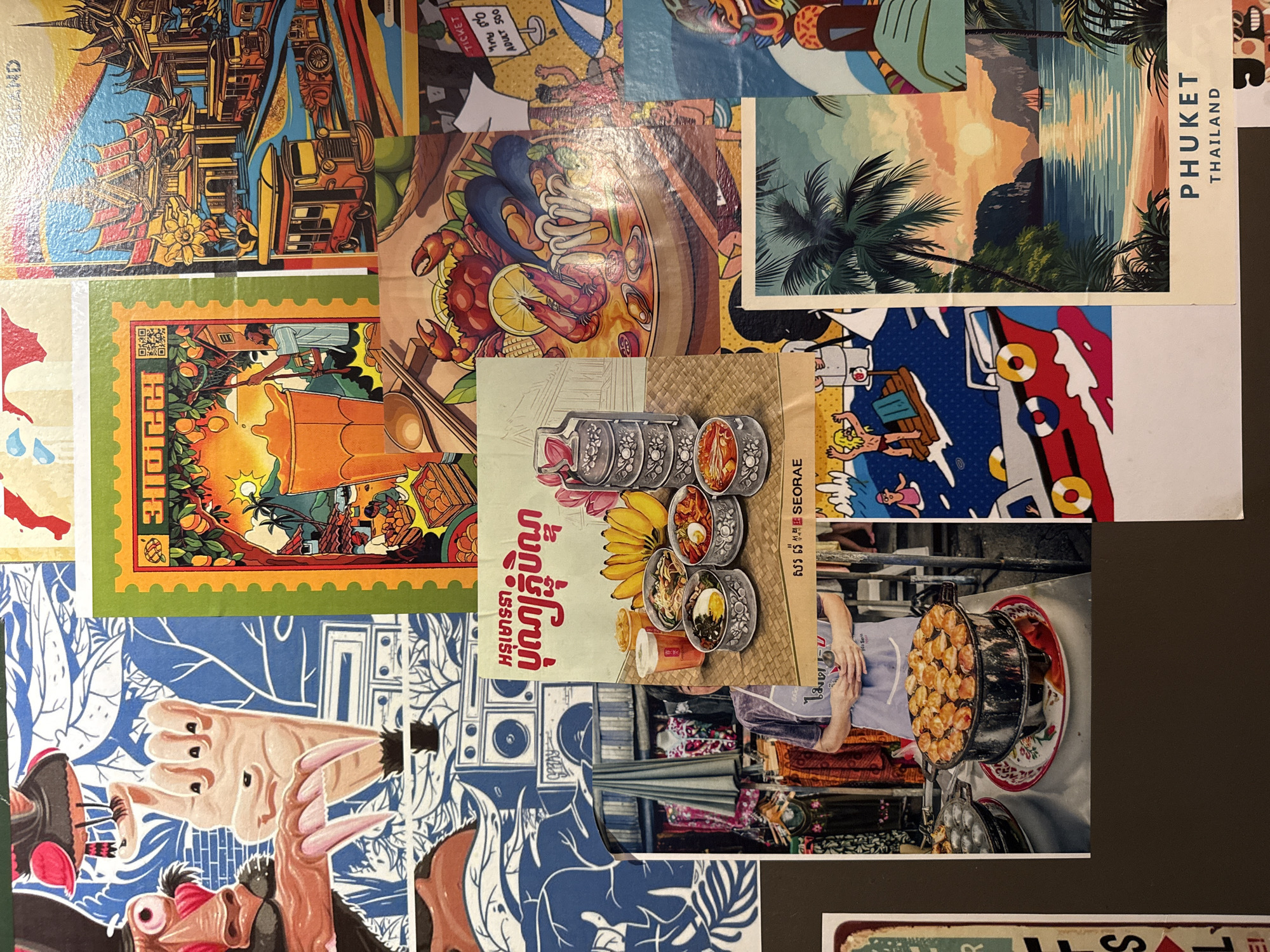 The image is a collage of various pieces of artwork and photographs, likely arranged on a surface. There is no visible indication of the city of Izhevsk, Russia, or any specific location within it.
The collage features several distinct elements:
* **Left side:** A large piece of artwork depicts a stylized, cartoonish face with large pink features and sharp claws, rendered in a blue and white pattern. To its right is a graphic of a hand with three extended fingers and claws.
* **Top center:** A green-bordered image resembles a stamp or postcard, featuring an illustration of a man harvesting fruit from a tree, a large glass of orange liquid, and a landscape with buildings and palm trees. Text in a non-Latin script is present at the top.
* **Top right:** A colorful illustration of ornate Asian-style buildings, with stylized buses and a roadway in the foreground. The text "THAILAND" is visible at the bottom right of this section.
* **Middle center:** A photograph shows a person wearing a purple apron, standing behind a stall with clothing and umbrellas displayed. In front of the person is a large, round cooking pot with numerous small, golden-brown circular items being cooked. A tiered metal food container is also visible to the left of the cooking scene, with small bowls of food. Text in a non-Latin script is present above this photograph.
* **Center right:** An illustration of a plate of seafood, including a crab, shrimp, mussels, and mushrooms, with a lemon slice.
* **Bottom right:** A postcard-style image of a tropical beach scene at sunset, with palm trees, a body of water, and a sailboat. The text "PHUKET THAILAND" is printed below this image.
* **Bottom center:** A portion of another artwork featuring a cartoonish character on a raft in blue water.
The overall impression is a collection of travel-related imagery, possibly souvenirs or promotional materials. The lighting suggests a bright environment, but the arrangement is flat, indicating it is likely a tabletop display or a wall collage.