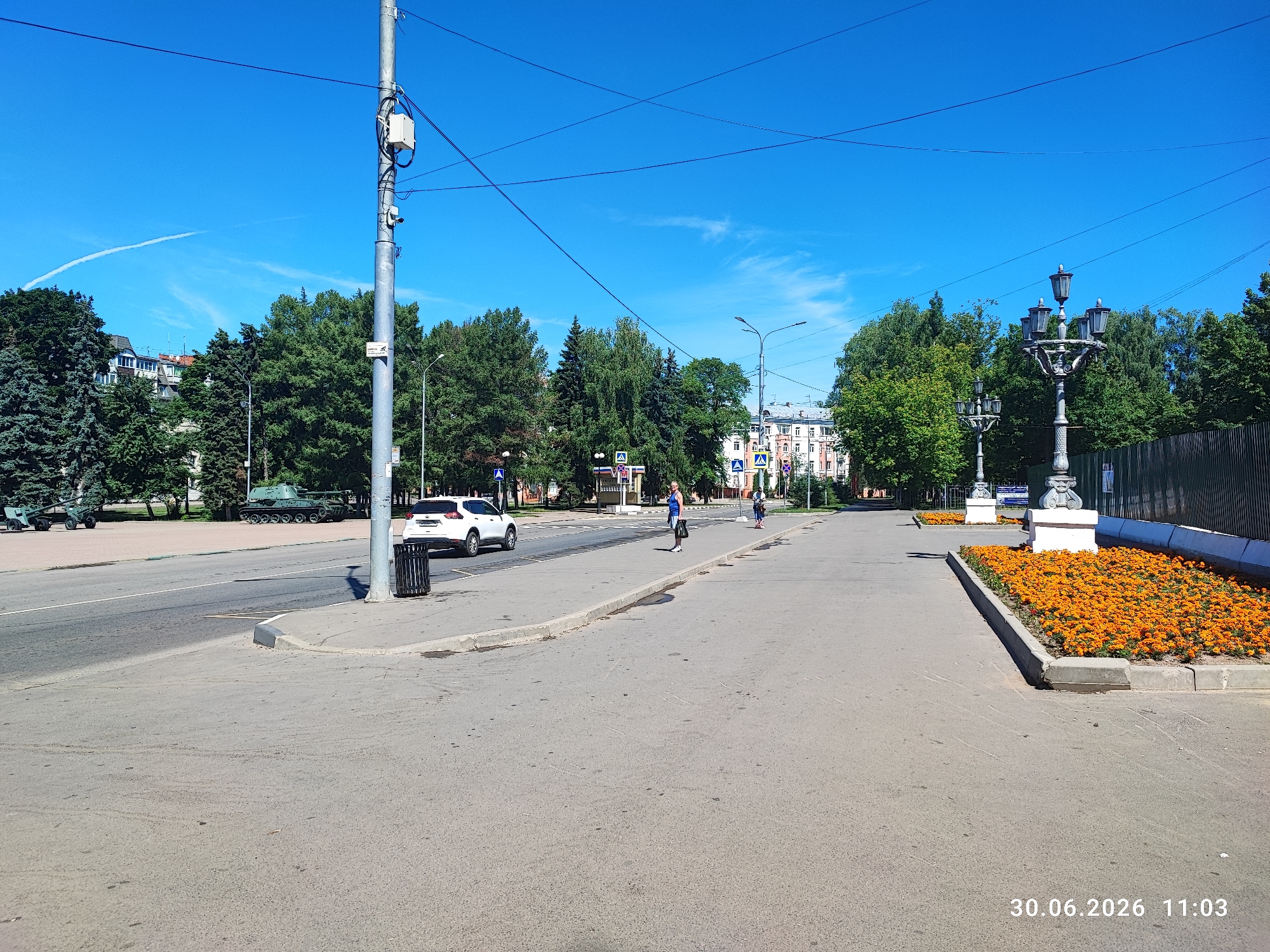 The image depicts a street scene in Lytkarino, Russia, with a clear blue sky overhead. In the foreground, there is an asphalt road and sidewalk. A white car is parked on the side of the road, and a trash can is situated on the sidewalk. Further down the street, a person is standing near a crosswalk, and another person is walking across the street. Several trees line the street, providing shade. In the background, there are buildings and a military tank on display. The date and time stamp on the image indicates "30.06.2026 11:03".