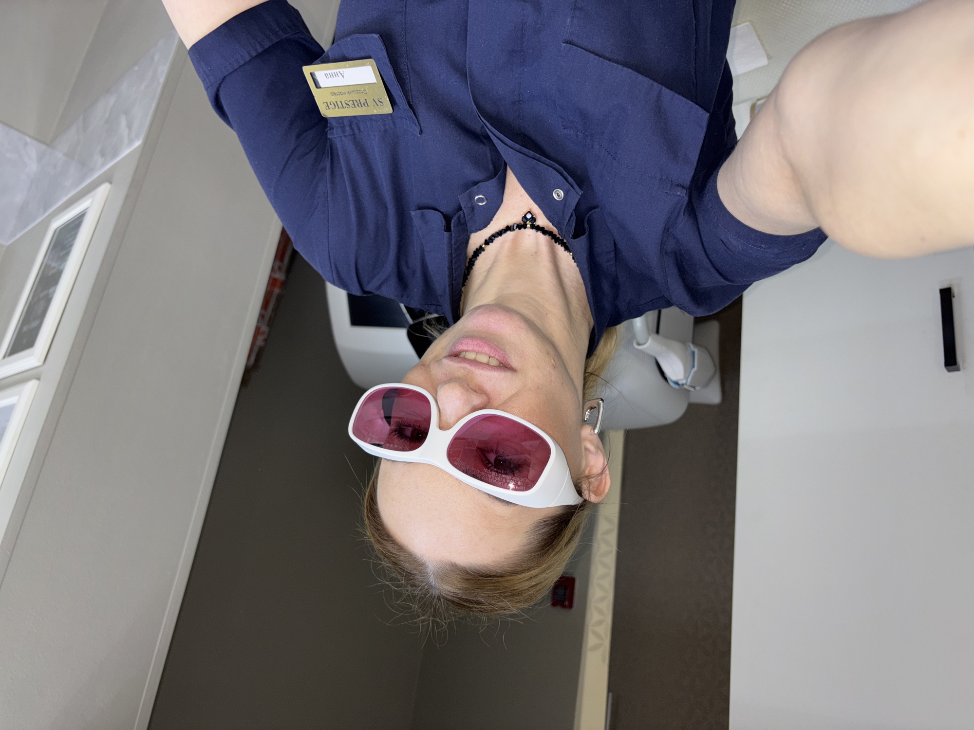 A selfie, taken by a fair-skinned woman with blonde hair, shows her wearing white-framed glasses with pink lenses. She is positioned upside down relative to the frame, wearing a dark blue button-down shirt with a name tag indicating "Anna" from "SV Prestige". She has a black beaded necklace. The location is identified as Vsevolozhsk, Russia.
Behind her is a white dental chair. A white wall with trim is visible on the left and a dark wall with a small red box mounted to it is on the right. The flooring is a neutral shade.