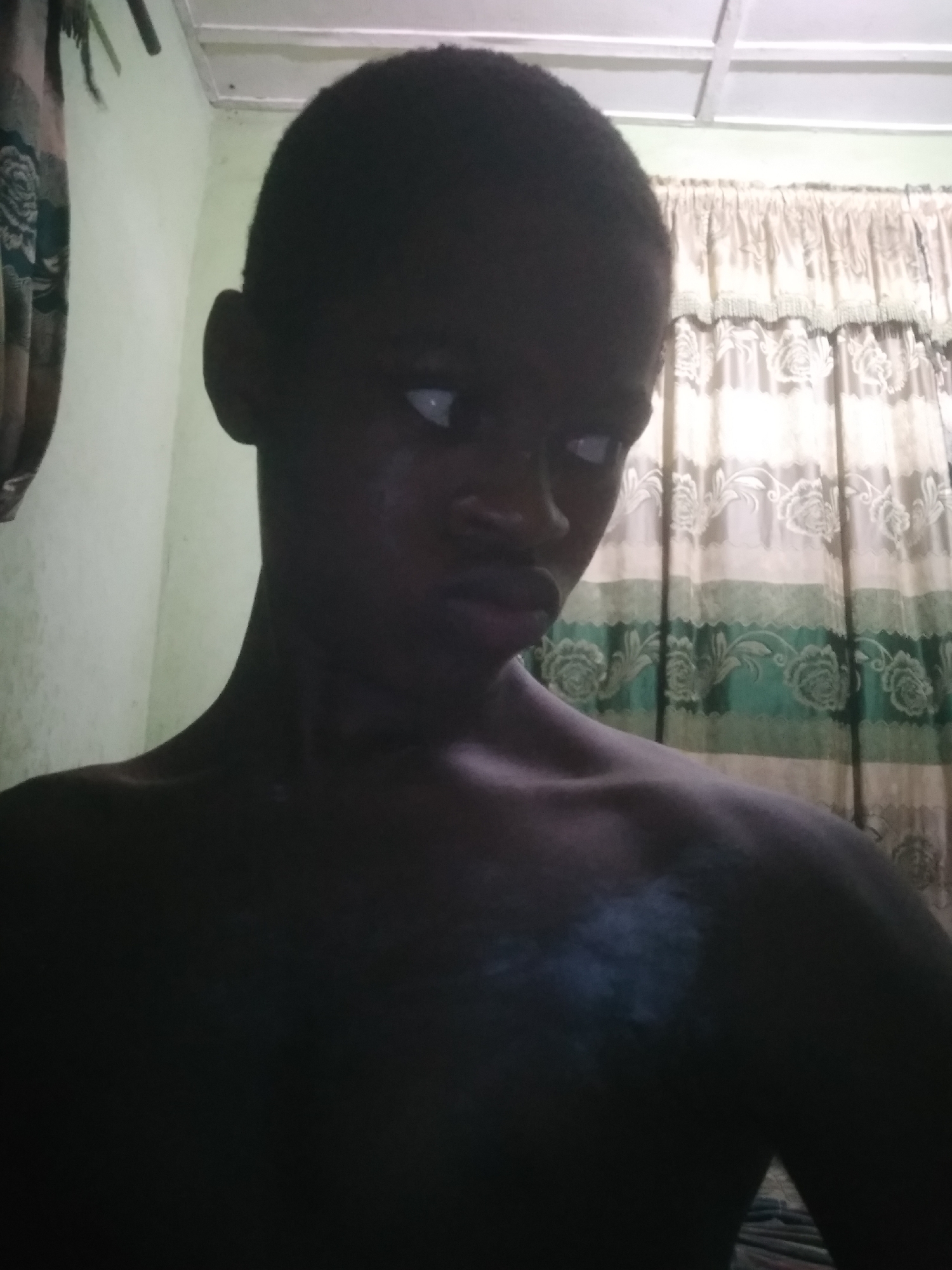 A medium shot depicts a shirtless young, dark-skinned individual looking to the right. The lighting is dim, obscuring details of the person's torso. Curtains with a floral pattern hang in the background. The location is Shangisha, Nigeria.