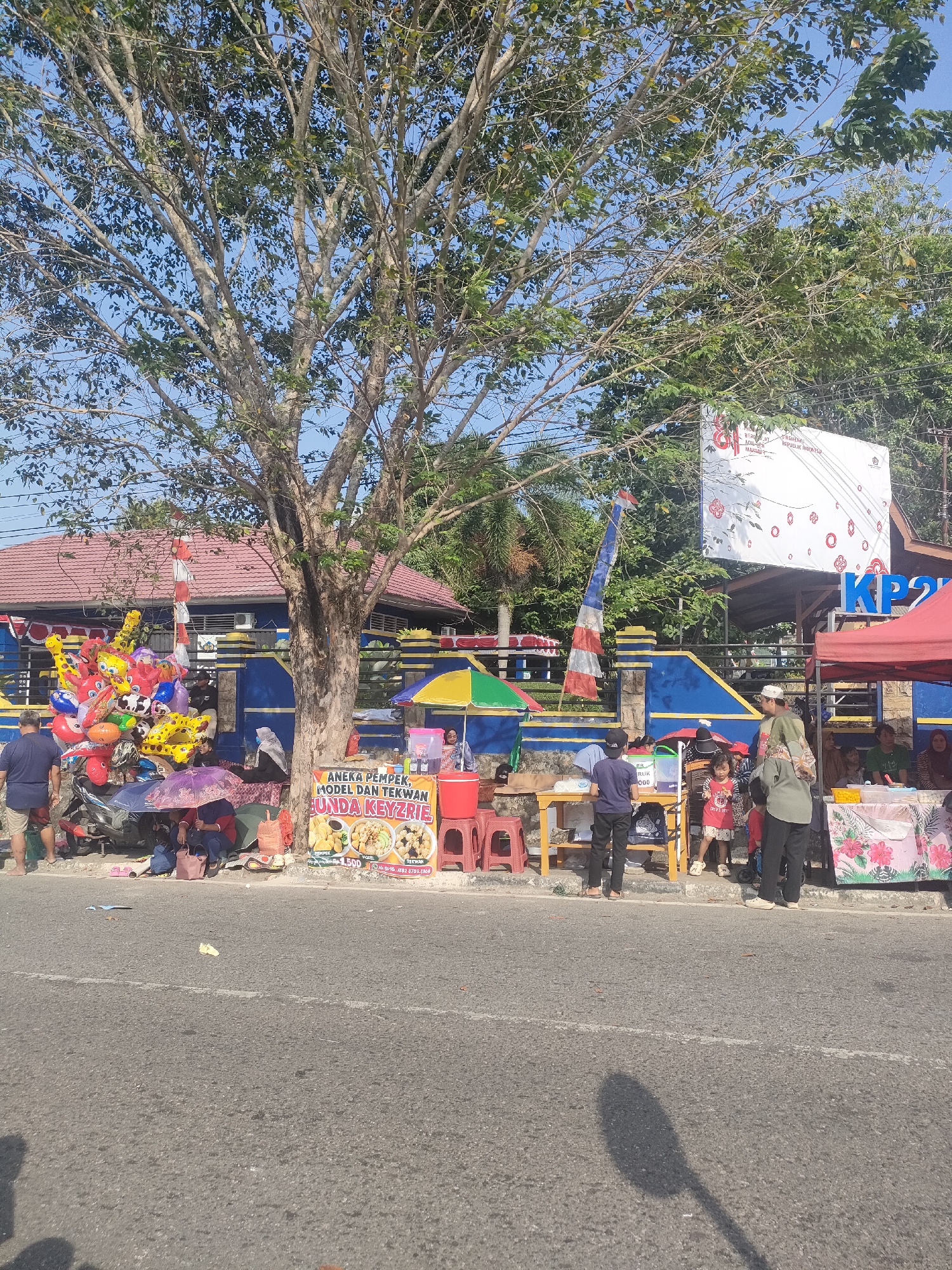 The image depicts a street scene in Sungailiat, Indonesia, likely a commercial area. In the foreground, a paved road runs horizontally across the frame, with the shadow of a streetlamp cast upon it. To the right of the road, a row of small roadside stalls operates under a bright, sunny sky.
The stalls are selling various goods, including what appears to be food and drinks, indicated by colorful signs and displays of products. One prominent sign advertises "ANEKA PEMPEK, MODEL DAN TEKWAN" (various pempek, model, and tekwan), suggesting local culinary offerings. People are seen interacting with the vendors, some standing, some sitting under umbrellas. Among the stalls, a large tree with sparse foliage provides some shade.
In the background, a blue building with a red-tiled roof serves as a backdrop to the stalls. A large white banner with red and white accents is mounted on a pole near the building. Flags, including an Indonesian flag, are also visible, adding a patriotic element to the scene. Several people are present, including vendors and customers. One stall displays a collection of colorful balloons, likely for sale. The overall impression is of a lively, informal marketplace on a typical day in Sungailiat.