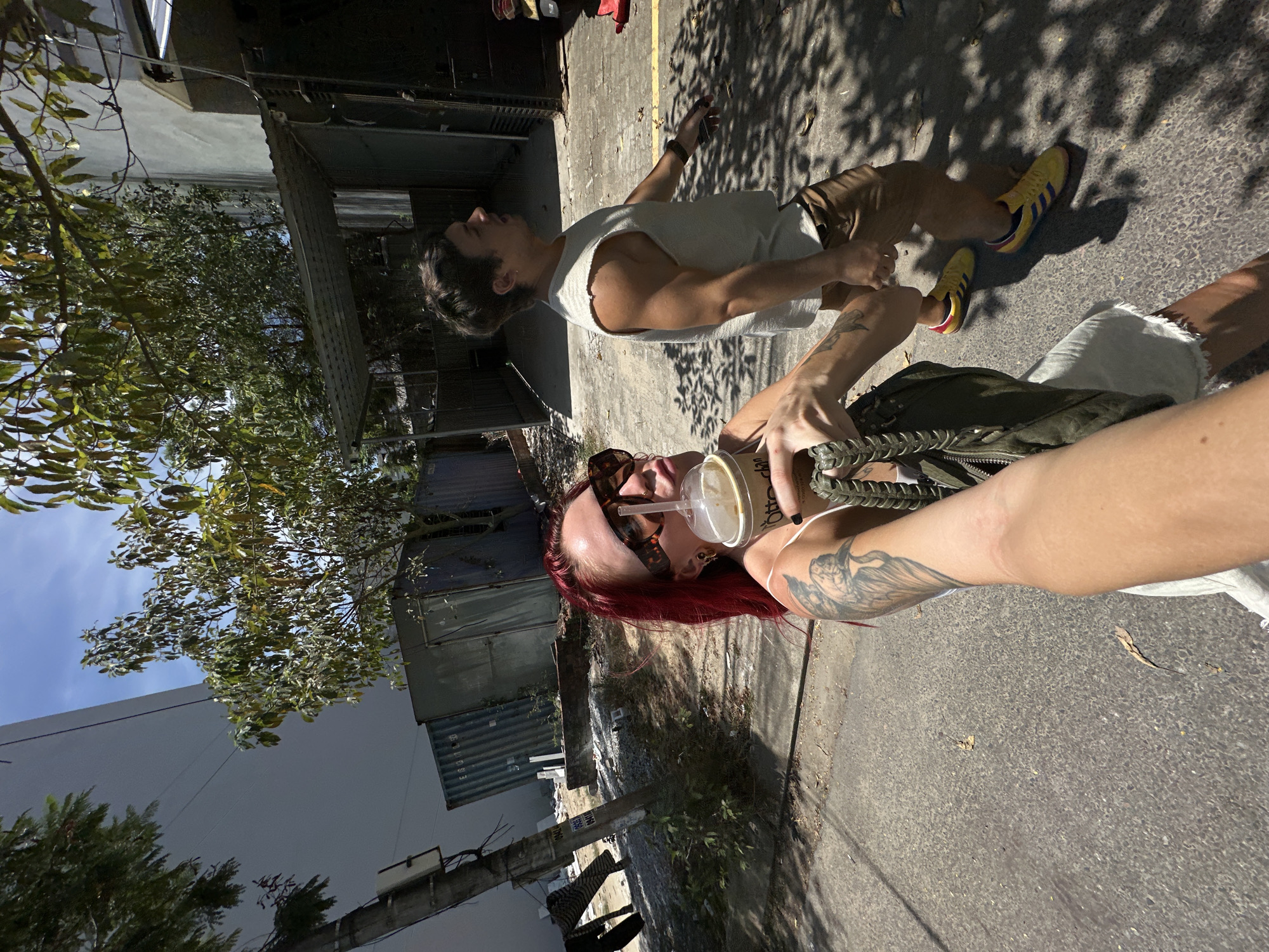 A woman with vibrant red hair and sunglasses holds a coffee cup while walking down a paved street in Đà Nẵng, Vietnam. She wears a white top and has tattoos on her arm. Behind her, a man in a light-colored tank top and brown shorts walks away from the camera. The street is lined with trees casting shadows on the ground, and there are shipping containers and a simple building in the background. The sky is clear blue with some clouds.