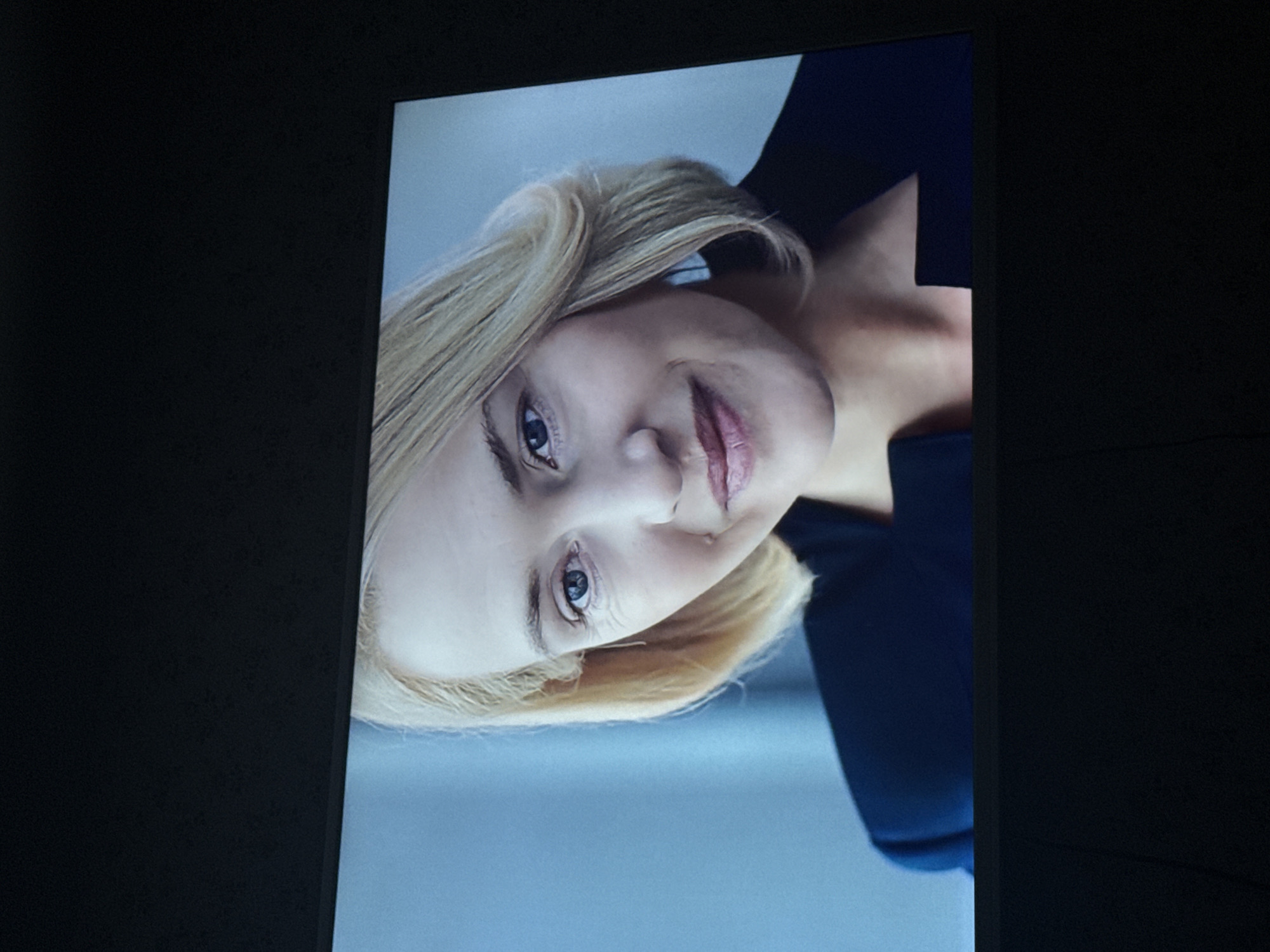 The image shows a screen displaying a portrait of a fair-skinned person with blond hair. The person is wearing a dark blue garment. The screen is surrounded by a dark, neutral-colored background. There is no information to suggest this image was taken in Mytishchi, Russia.