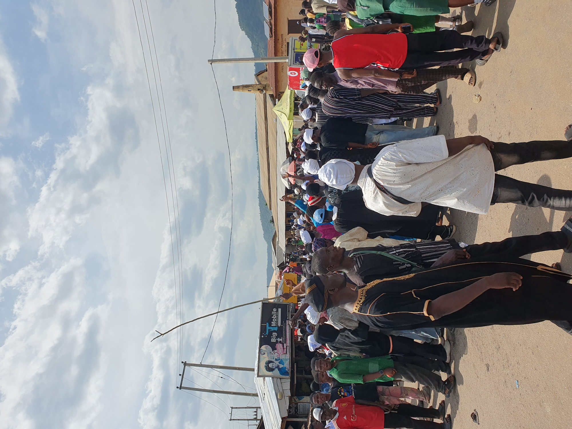 A daytime scene showing a large group of people gathered on a street in Lokoja, Nigeria. The crowd is diverse in terms of clothing, with many wearing traditional attire, including white caps, gowns, and colorful fabrics. Some individuals are wearing more casual clothing like t-shirts and jeans. The gathering appears to be densely packed along a paved road.
In the background, there are buildings, utility poles, and electrical wires. A sign with the words "BIG Mobile" is visible on a building or structure next to the crowd. The sky is partly cloudy with light blue patches. The surrounding landscape includes a mountain range in the distance, suggesting the scene is in a more urban or semi-urban setting.