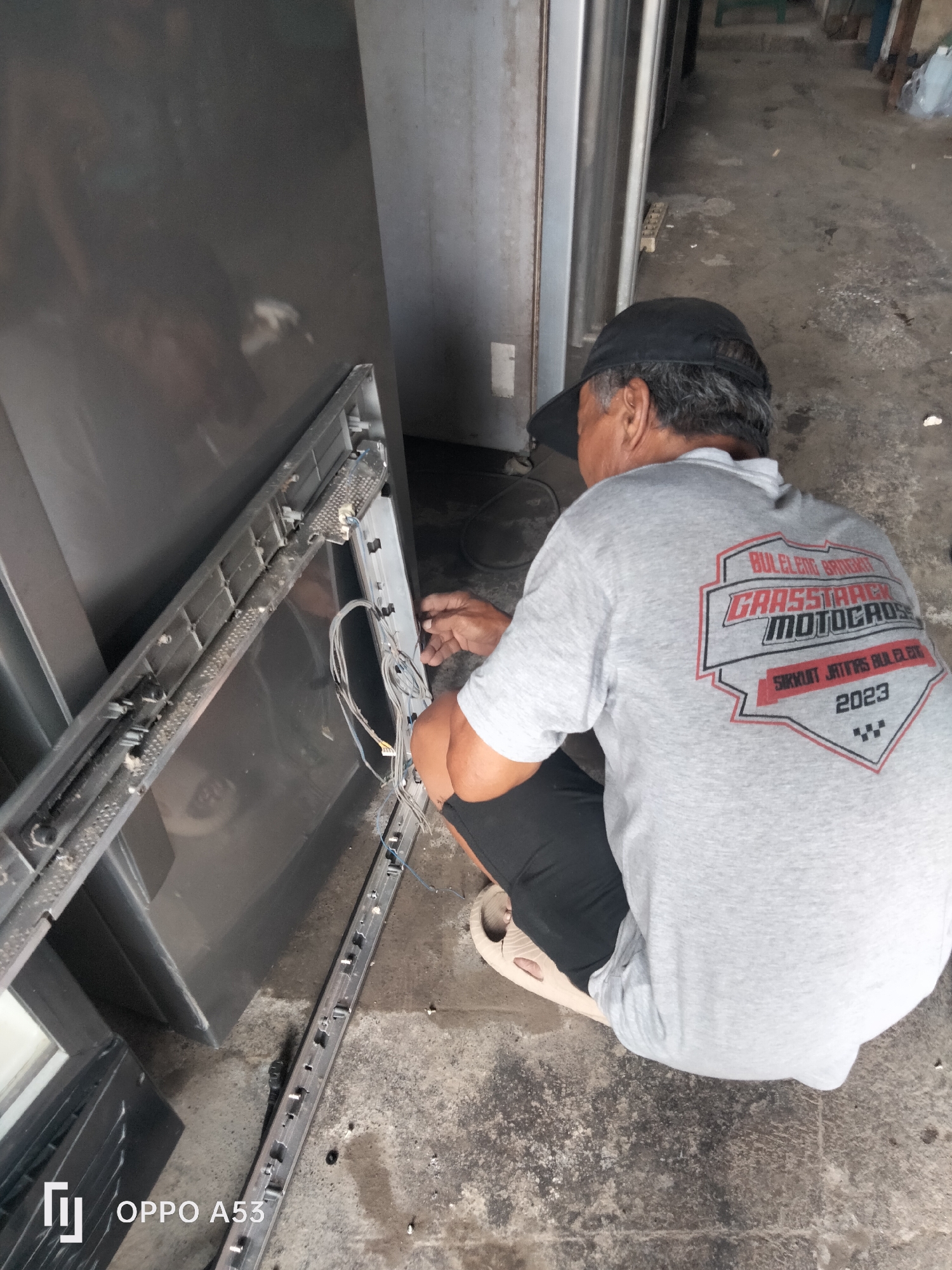 In Denpasar, Indonesia, a man wearing a gray t-shirt with "GRASSTRACK MOTOCROSS" and "2023" printed on the back, along with a black baseball cap, is working on what appears to be a broken appliance, possibly a refrigerator. He is kneeling on a concrete floor, with the appliance disassembled and its internal components exposed, including wires and metal tracks. The environment is outdoors, with a rough concrete floor and what looks like a large, dark gray metallic structure, possibly another appliance or building panel, to the left. The overall scene suggests a repair or dismantling operation in an urban or industrial setting.