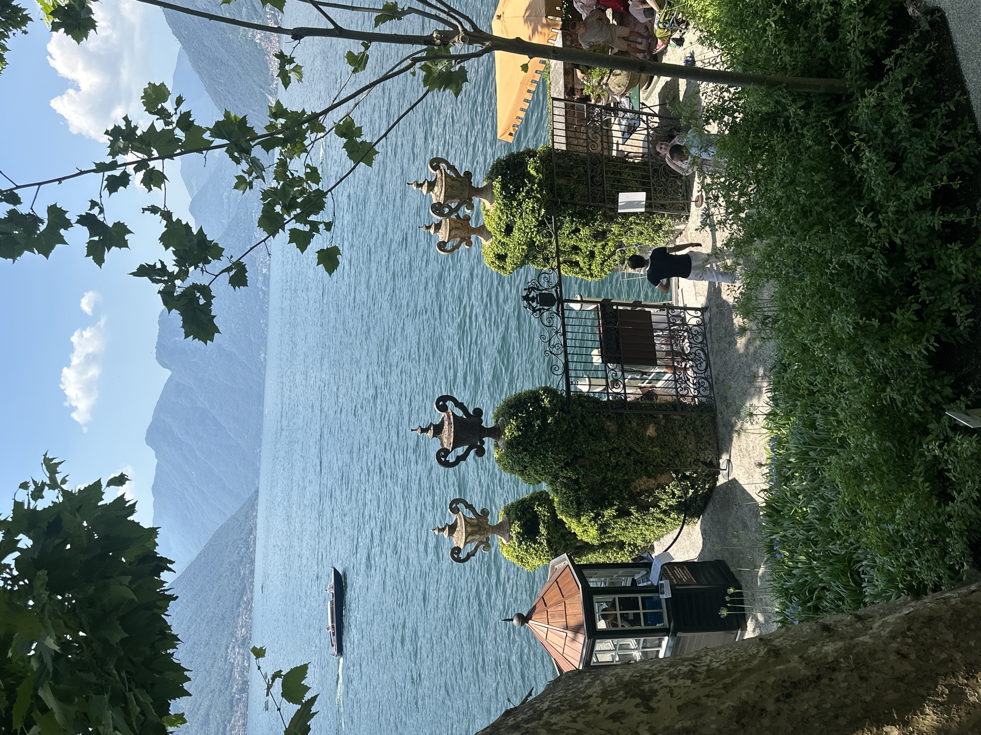 The image depicts a scenic view of Lake Como in Tremezzina, Italy. In the foreground, lush green foliage frames the scene, with a large tree trunk on the left and branches overhead. A small, ornate gazebo with a copper roof is visible on the left, beside a black structure with windows, possibly a ticket booth or information kiosk.
Dominating the middle ground are two elaborately trimmed topiaries shaped like urns, each topped with a decorative urn statue. Between them is an open wrought-iron gate, with more ornate fencing to the right. Beyond the gate, a paved area is visible, with a few people seated at tables under a yellow umbrella.
The water of Lake Como stretches into the background, with a boat cruising across its surface. In the distance, rolling mountains rise, covered in verdant vegetation and dotted with buildings, suggesting the presence of villages along the shore. The sky is a clear blue with a few wispy clouds, indicative of a sunny day.
Several people are present in the image. A man in a black t-shirt and white pants stands near the gate, facing away from the camera. A woman and a child are visible in the background, near the seated patrons. Another person is partially visible inside the gazebo. The overall scene conveys a serene and picturesque atmosphere of a lakeside garden estate.