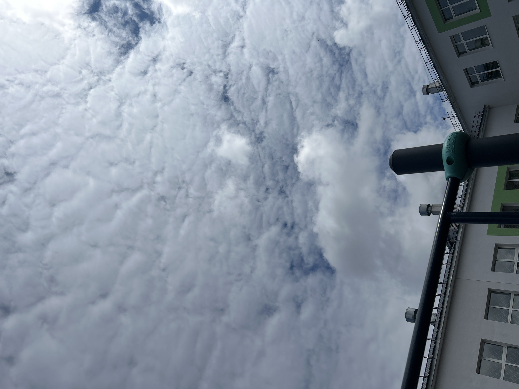 A low-angle shot, likely from a person standing on the ground in Chaikovsky, Russia, presents a view of the sky filled with a dense layer of altocumulus clouds. Patches of blue sky are visible between the cloud formations. The perspective includes part of a building with white walls, green window trim, and several windows with grid patterns. A section of a black metal structure, possibly a balcony or some other architectural element, is visible in the foreground. A cylindrical object with a light green brace is attached to the metal structure. The photograph's orientation suggests it was taken looking upwards.