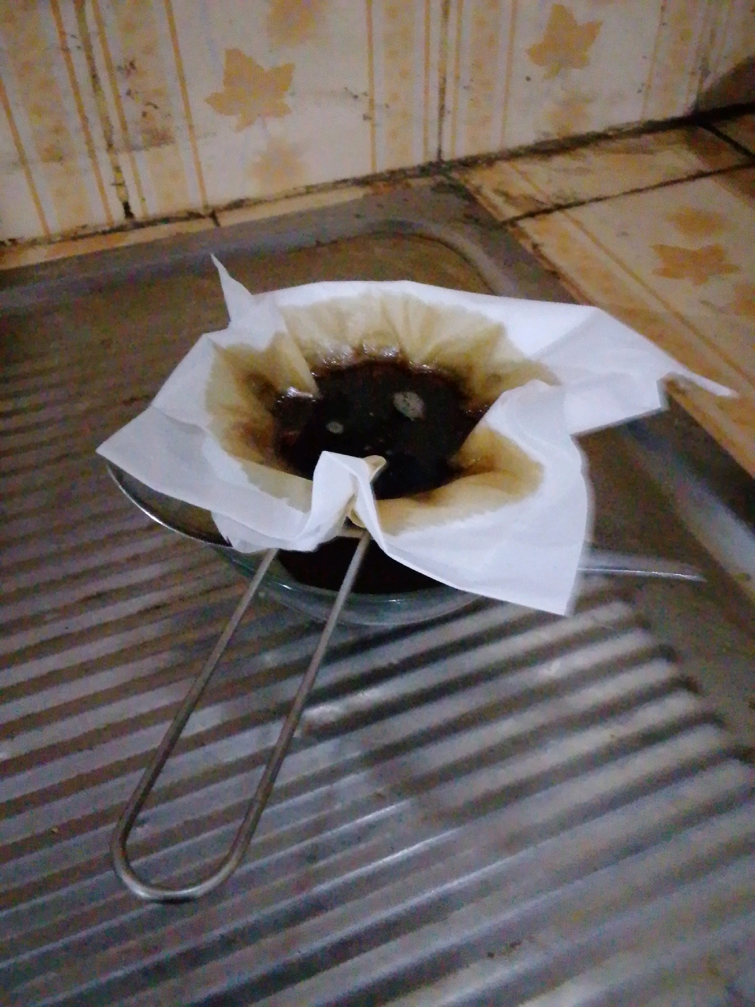 A close-up shot shows a stainless steel sieve with a paper coffee filter filled with dark brown liquid and coffee grounds, resting over a stainless steel sink. The filter is wet and stained with coffee. The sink has visible ridges. The background features a tiled wall with a light yellow and white striped pattern and faded leaf imprints. The scene appears to be in a kitchen or utility area, possibly in Drancang, Indonesia, given the context. There are no people visible in the image. The lighting is dim, and the image quality is grainy.