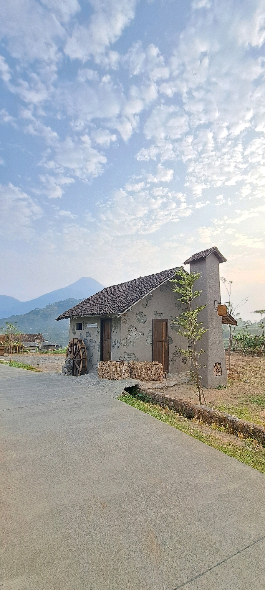 The image depicts a rustic, stone-clad building with a pitched, tiled roof, situated in a rural landscape. The building has two wooden doors and a prominent stone chimney on the right side. Adjacent to the building, on the left, is a wooden water wheel supported by stones. Two bales of straw are placed in front of the doors. A small, leafy tree grows to the right of the chimney. In the background, rolling hills and a distant mountain are visible under a sky filled with scattered white clouds. The foreground is dominated by a paved road or pathway. The scene is set in the village of Tamiajeng, Indonesia, characterized by its natural beauty and traditional architecture. The lighting suggests it is either early morning or late afternoon, with soft sunlight illuminating the scene. No people are present in the image.