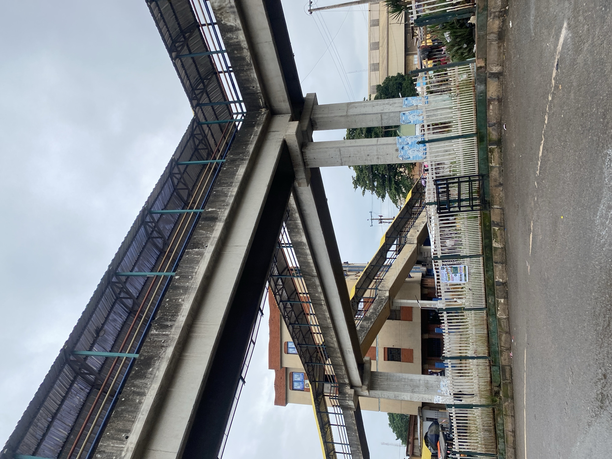 An elevated pedestrian walkway, likely part of a transportation hub or an overpass, is prominently featured in the image, shot from a low angle on a street. The walkway is constructed of concrete and has a corrugated metal roof supported by a network of metal beams and poles. The roof is in a weathered condition, showing signs of age and exposure to the elements. The walkway is supported by large concrete pillars.

Below the walkway, a building with light-colored walls and darker brick accents is visible. It has several windows, some of which appear to be blue. Attached to the building are ramps leading to the elevated walkway, also with yellow railings. A white picket fence runs along the front of the property, separating it from the street. Behind the fence, there are signs and some posters on the concrete pillars. Some of these posters feature portraits of individuals.

The street in the foreground is paved and appears to be wet or recently rained on, with a few white lines marking the traffic lanes. The sky is overcast and cloudy, suggesting a dull or rainy day. Trees are visible in the background, as are other buildings and structures, including what looks like a factory or warehouse with a light-colored, sloped roof. Electrical poles and wires are also present. The scene appears to be an urban environment in Abeokuta, Nigeria, characterized by functional, albeit somewhat aged, public infrastructure. There are no clearly identifiable people in sharp focus, though there are indistinct figures visible behind the fence and near the building. The overall impression is one of a busy, working urban area.