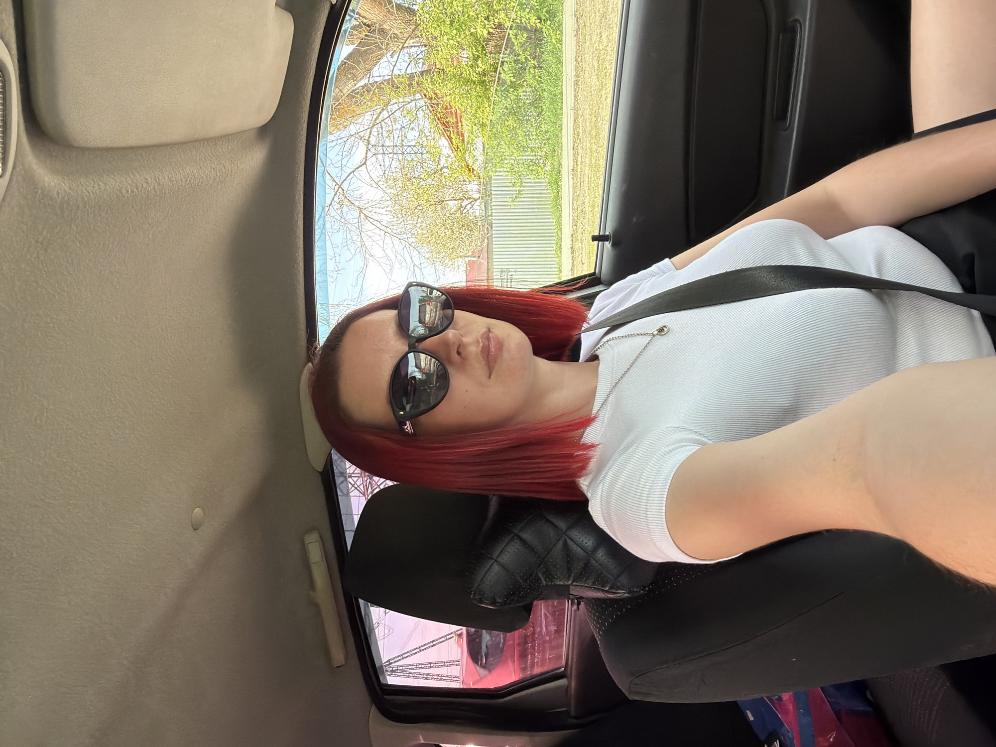 The image shows a person with red hair wearing sunglasses and a white ribbed shirt, secured with a seatbelt, sitting in a car. The background visible through the car window includes trees, a fence, and a building with a red roof. The interior of the car is predominantly grey and black. It is not possible to identify the specific location within Chaikovsky, Russia from the image.