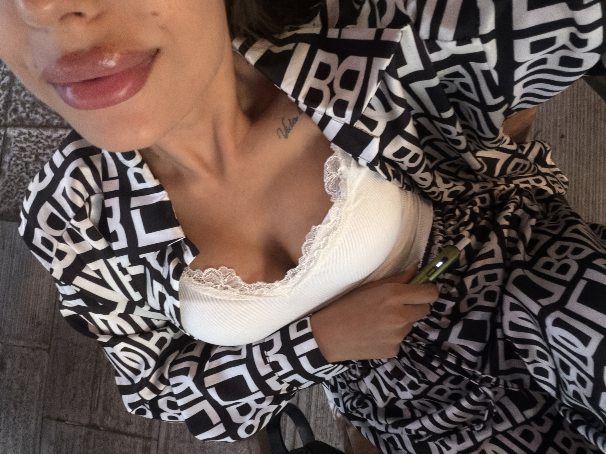 The image, taken in Svishtov, Bulgaria, is a close-up shot of a woman from the chest up. She is wearing a white ribbed top with lace trim and a black and white patterned jacket. The pattern on the jacket appears to be a repeating logo or abstract design. The woman is leaning back, with her tongue slightly out. A tattoo is visible on her collarbone. She is holding a small, green electronic device in her right hand, which is tucked into the waistband of her patterned skirt. The background is a textured surface, possibly a floor or a tiled area, with a slightly blurred appearance. The lighting suggests an outdoor or well-lit indoor setting.