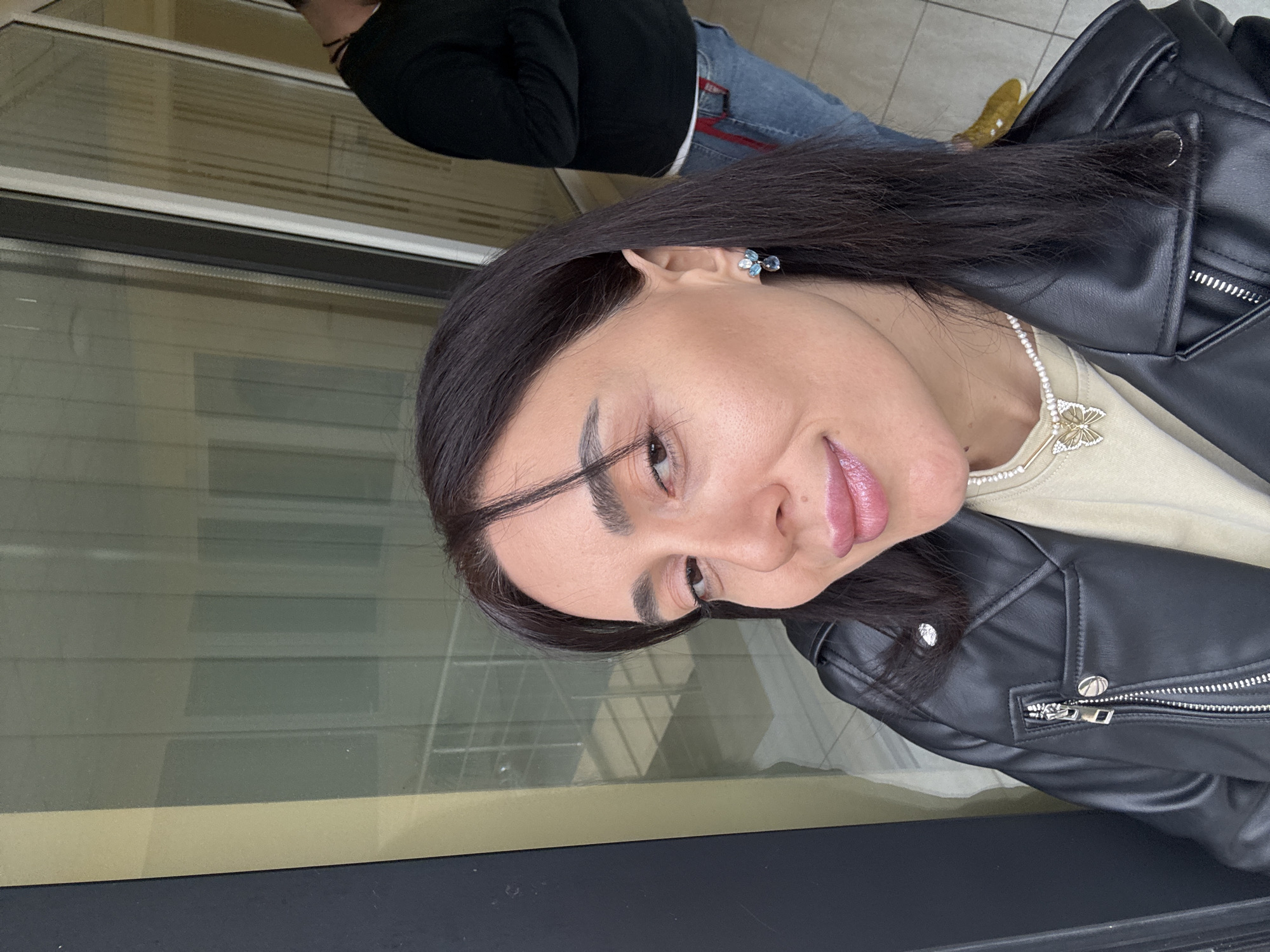 An image captures a woman in a selfie orientation, positioned in the foreground and slightly to the left, within the interior of a modern building in Tbilisi, Georgia. She has dark hair, brown eyes, and is wearing a black leather jacket over a light-colored top. A pearl necklace with a gold butterfly pendant is visible, as is a multi-stone blue earring in her right ear. Her expression is a subtle smile directed towards the camera. In the background, to the right, a man is partially visible from the chest down, wearing a dark long-sleeved shirt, blue jeans with a red accent on a belt loop, and yellow shoes. His back is mostly turned towards the camera. The background consists of large glass panels, reflecting and revealing elements of a building's interior, specifically a staircase with metallic railings. The setting appears to be a well-lit, possibly commercial or public, indoor space.