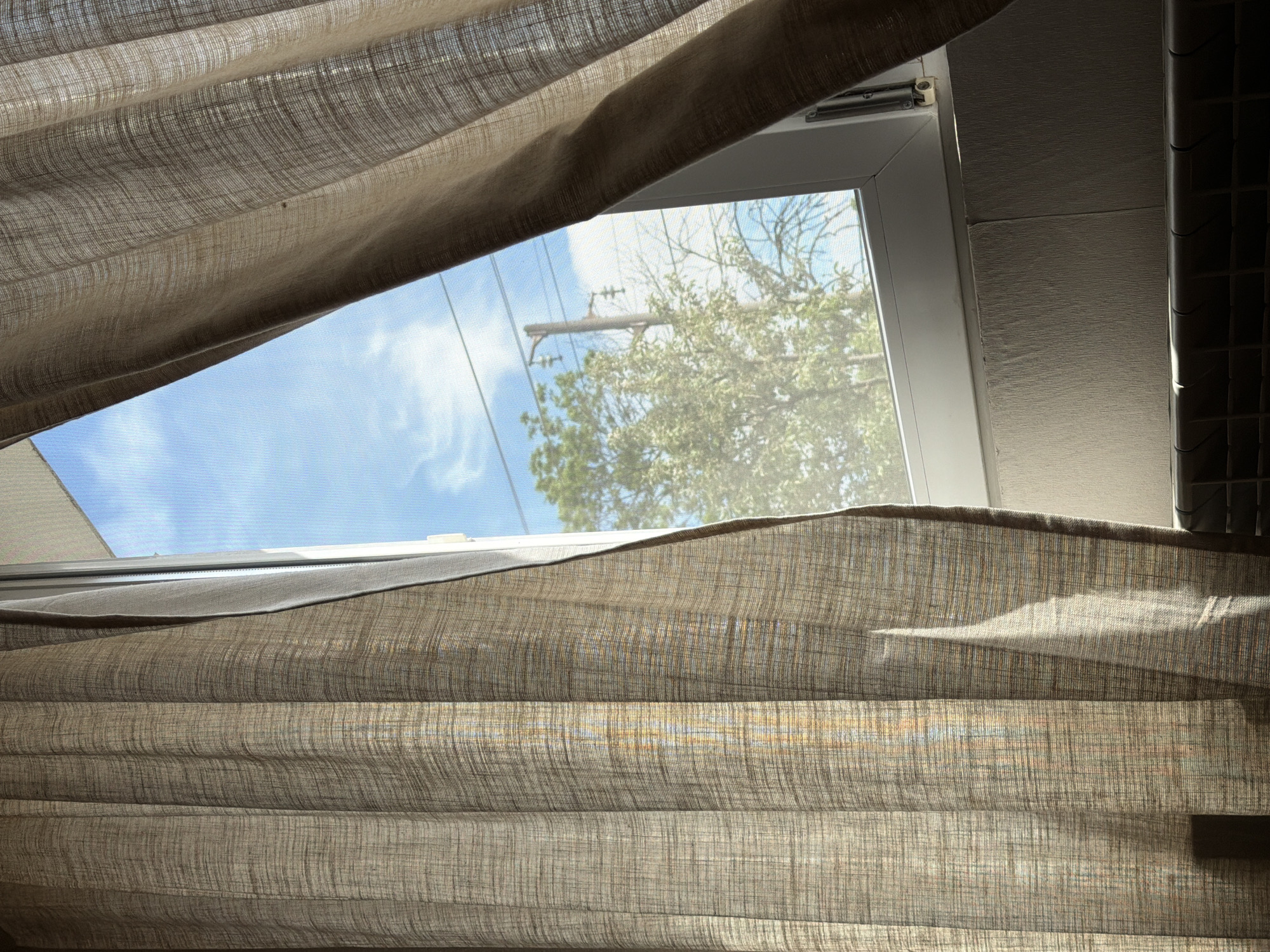 A partial view through a window partially obscured by a beige-colored fabric curtain. Outside, a view of a blue sky with scattered clouds and a tree with dense green foliage is visible. A utility pole with wires is also visible through the window. The window frame is white. The window is located in an interior space. There is no information available to determine that this is Selyanin, Bulgaria.