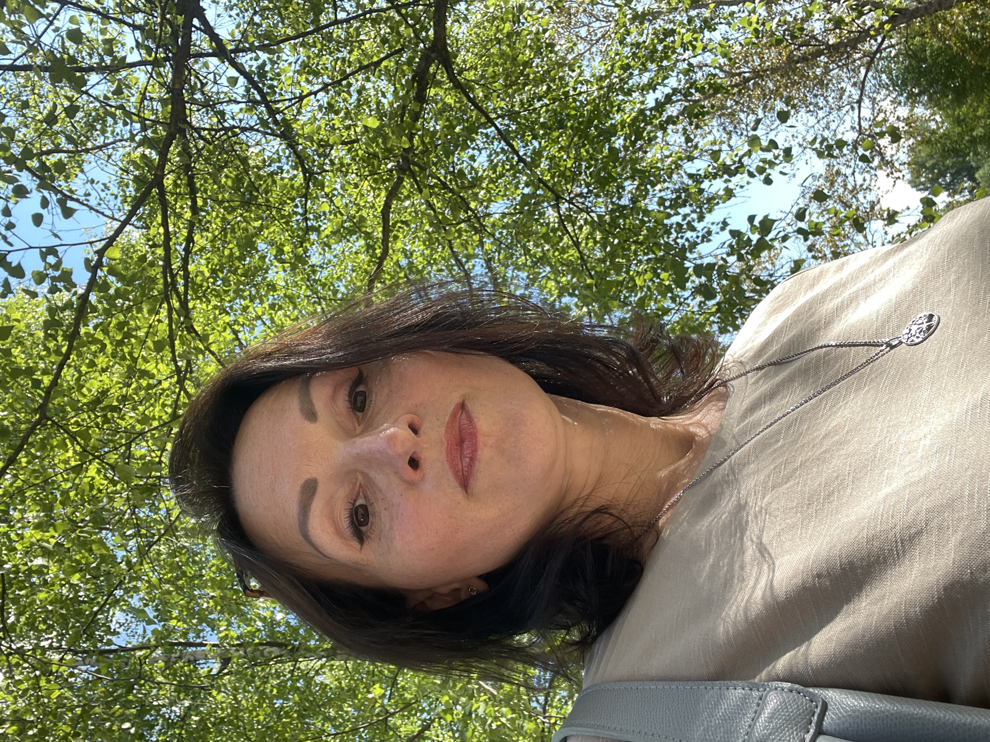 A medium shot, eye-level, outdoor selfie shows a middle-aged woman in the foreground, looking directly at the camera. She has dark, shoulder-length hair, brown eyes, and is wearing light-colored clothing with a silver necklace. She appears to be in a park or natural setting, with lush green trees and a blue sky visible behind her. The lighting suggests it is daytime. The specific location in Kirovo-Chepetsk, Russia, is not discernible from the image.