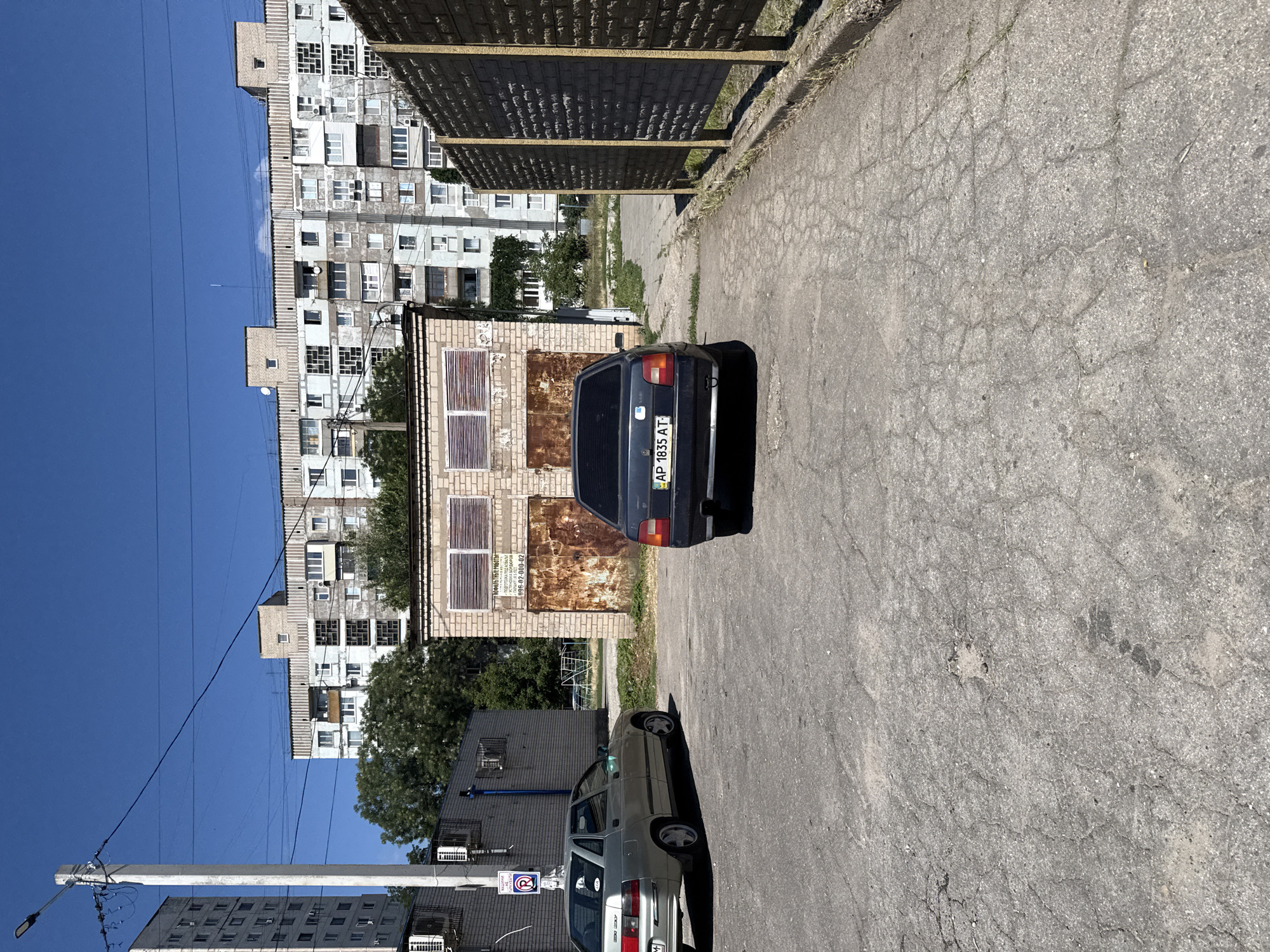 The image shows a parking area in front of a brick garage with rusted doors and gridded windows. A dark-colored sedan with license plate AP 1835 AT is parked in the foreground, facing away from the camera. To the left, a lighter-colored sedan is also parked. Behind the garage, a multi-story residential building with balconies and air conditioning units is visible. The sky is clear and blue. A street light stands to the left of the garage, and a grey brick wall with yellow posts runs along the right side of the frame. The ground is paved with cracked asphalt. This scene is located in the city of Zaporizhzhia, Ukraine.