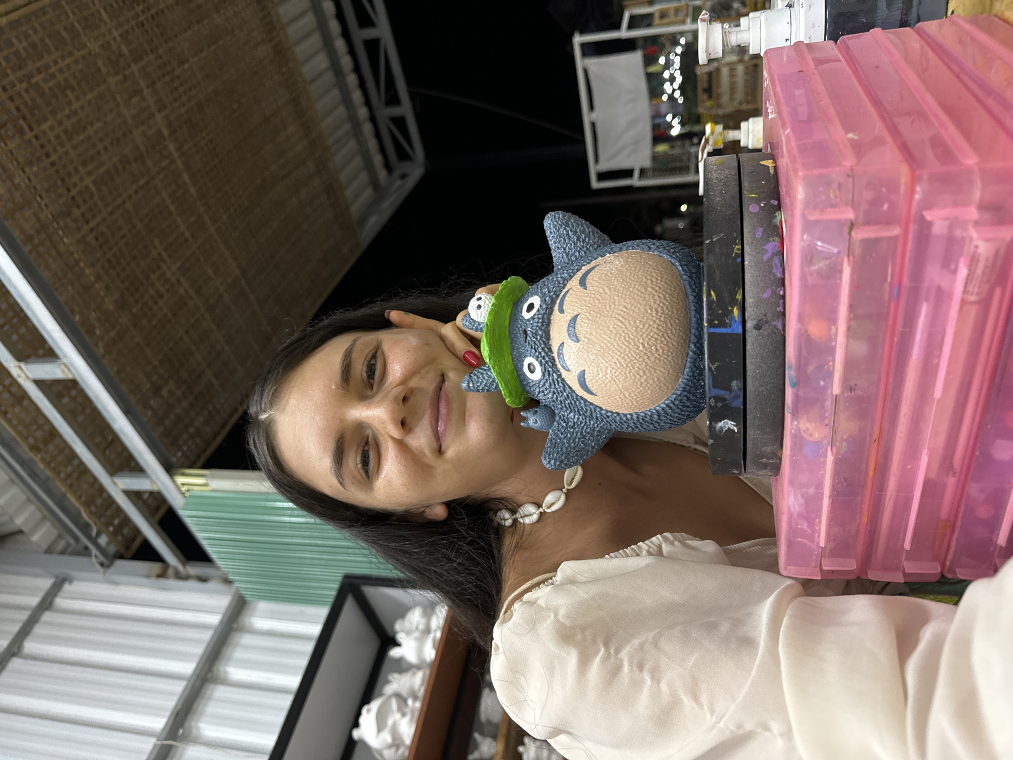 A young woman with long dark hair and a shell necklace smiles at the camera, her left hand resting on her cheek. She is wearing a cream-colored puffed-sleeve top. To her right, she holds a blue Totoro figurine with a green hat, which is perched on a stack of pink plastic containers. The background suggests an outdoor setting, possibly a workshop or craft area, with corrugated metal walls, wooden beams, and shelves with white ceramic objects. The location is identified as Mũi Né, Vietnam. The lighting indicates it is evening or night.